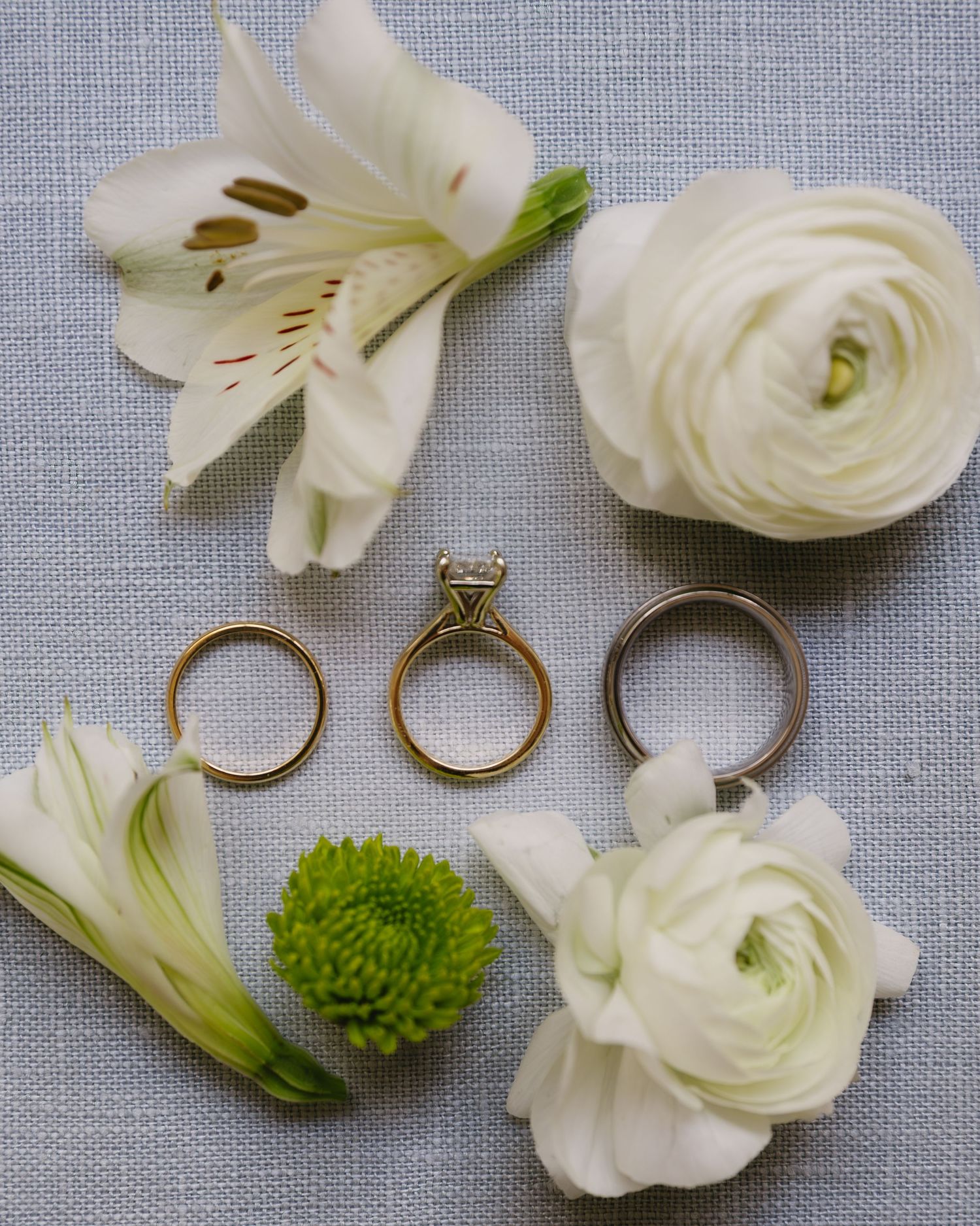 White ranunculus flowers and wedding rings arranged on light blue linen fabric.