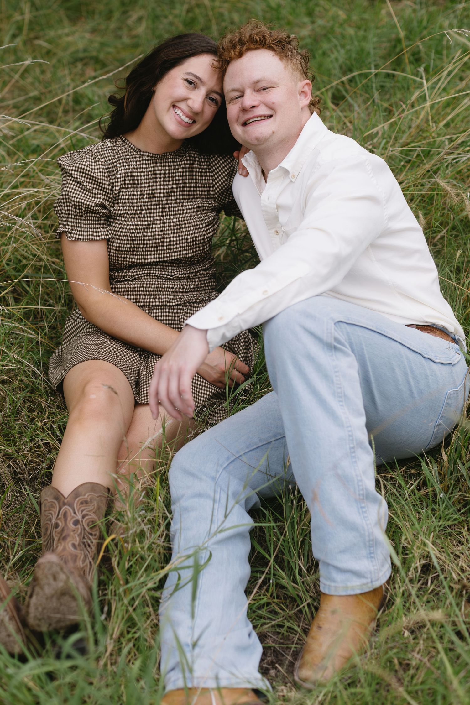 Couple wearing cowboy boots cuddles together on grassy ground during outdoor photoshoot.