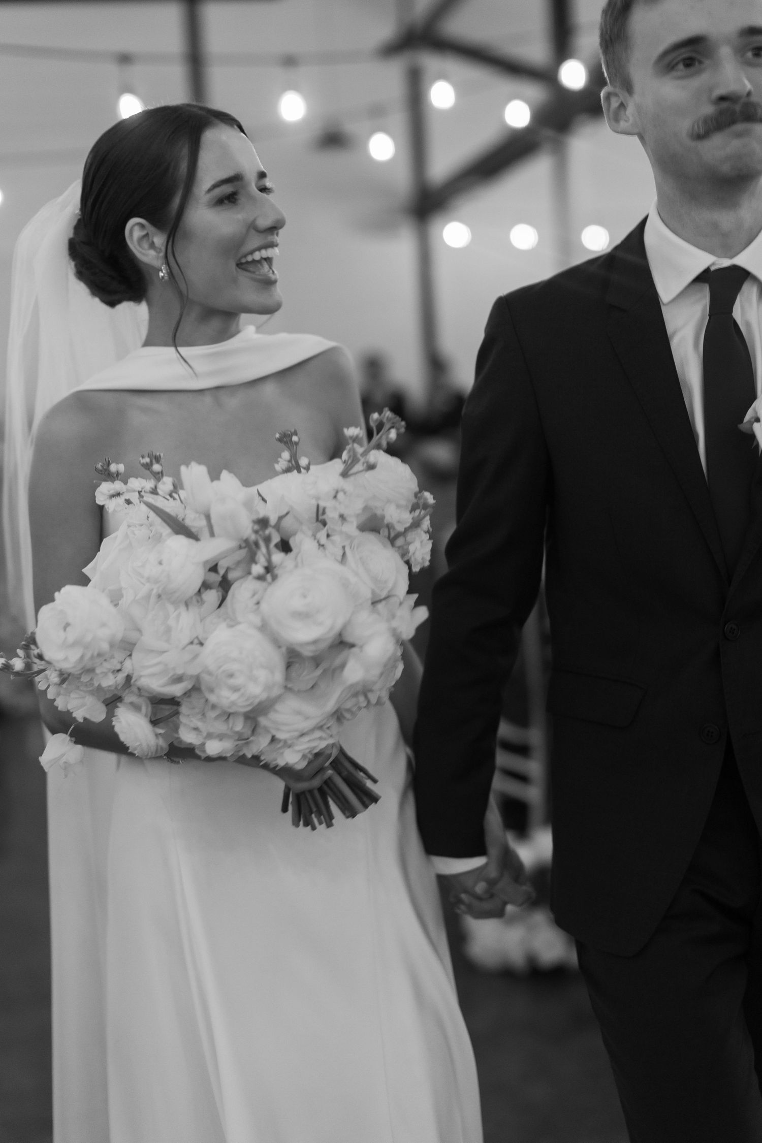 Black and white wedding photo showing elegant couple with white floral bouquet under twinkling lights.