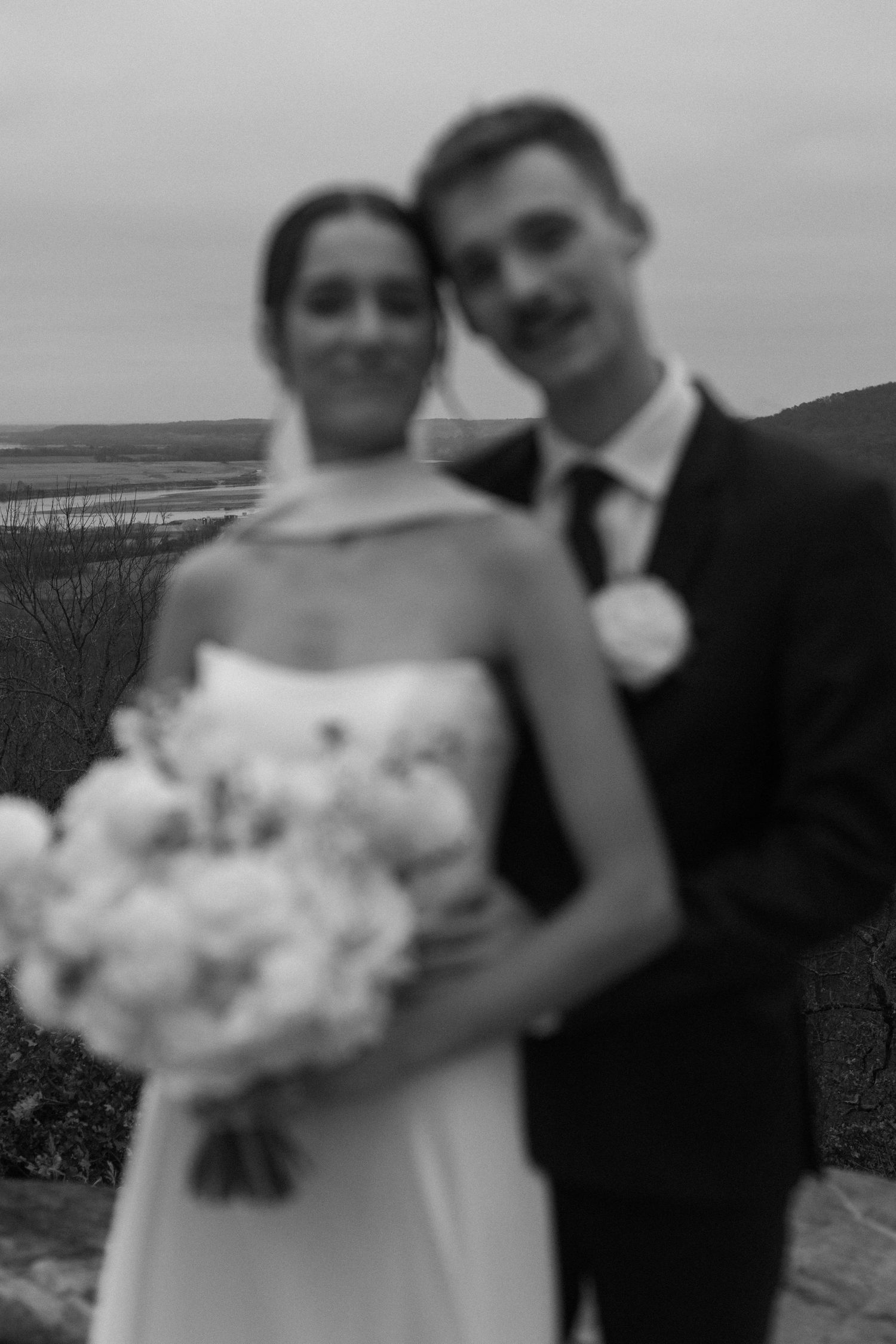 A soft focus portrait of newlyweds outdoors with a white bouquet against a cloudy sky.