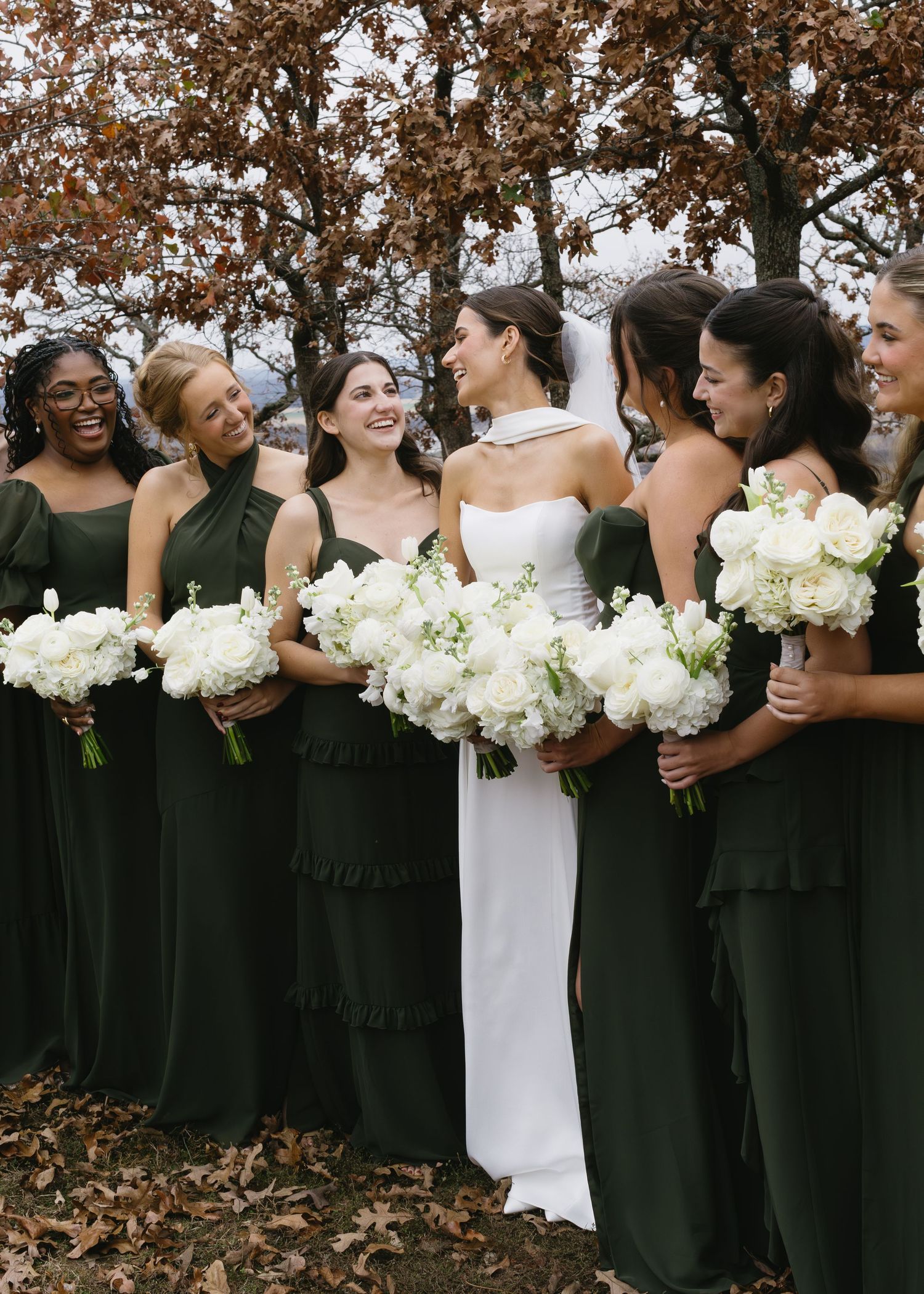 A bridal party in dark green dresses poses with white bouquets against autumn foliage.
