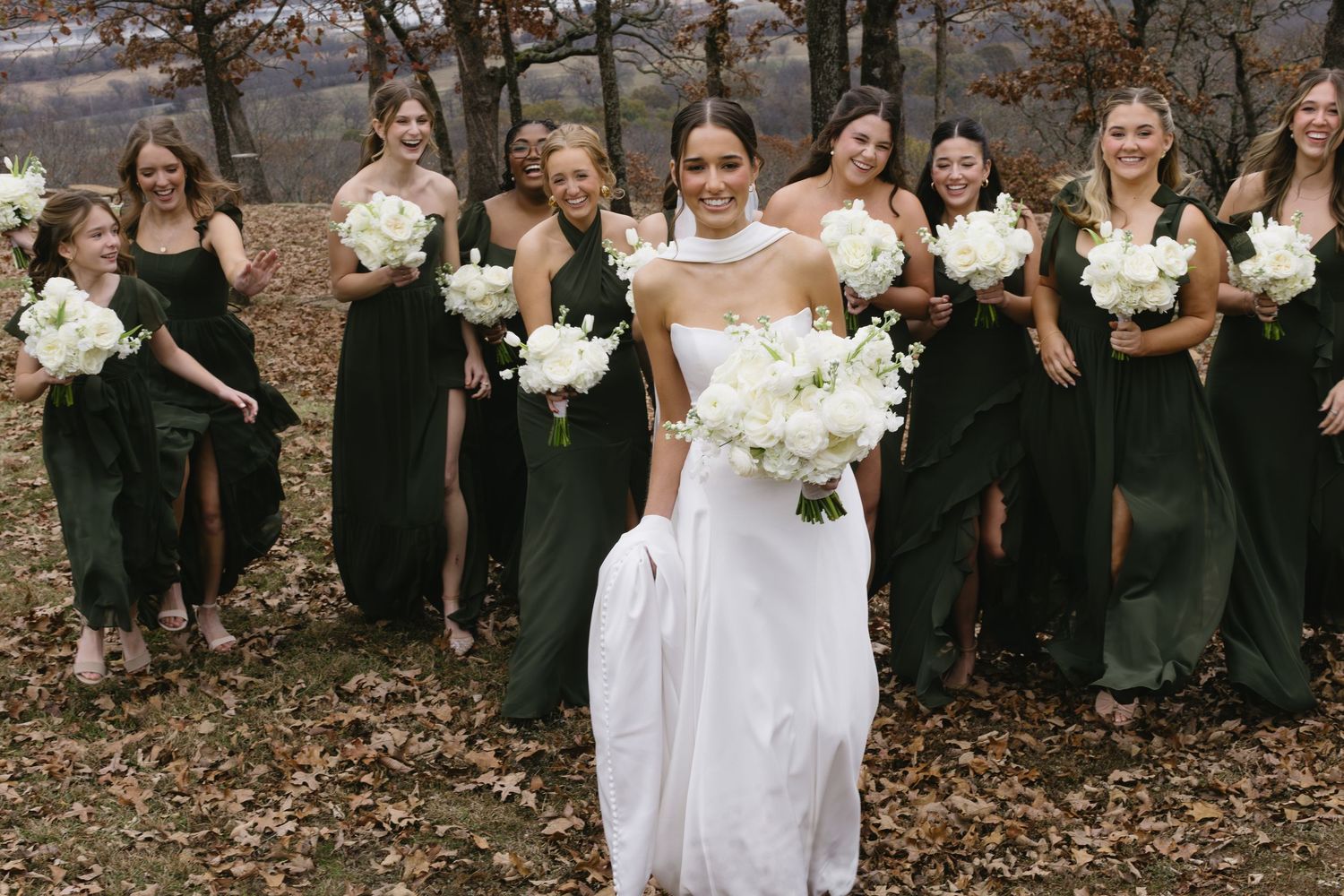 Wedding party in emerald dresses stands behind bride in white holding matching white bouquets.