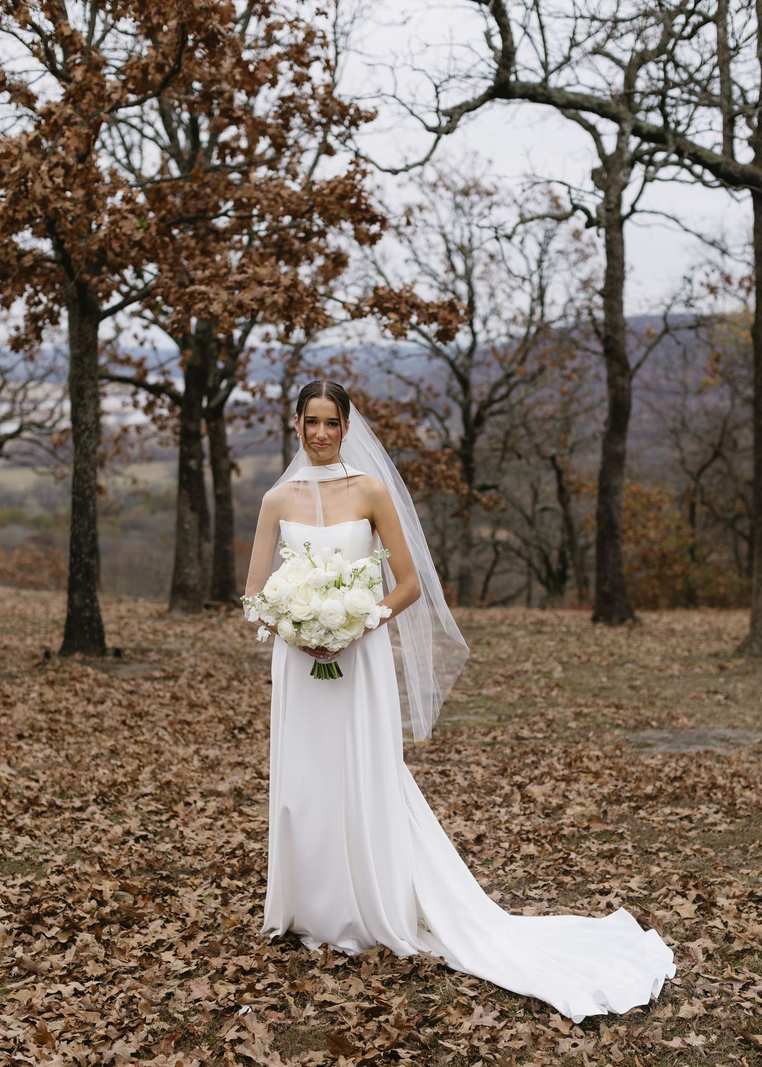 Bride in flowing white gown holding bouquet amid autumn trees and fallen leaves.