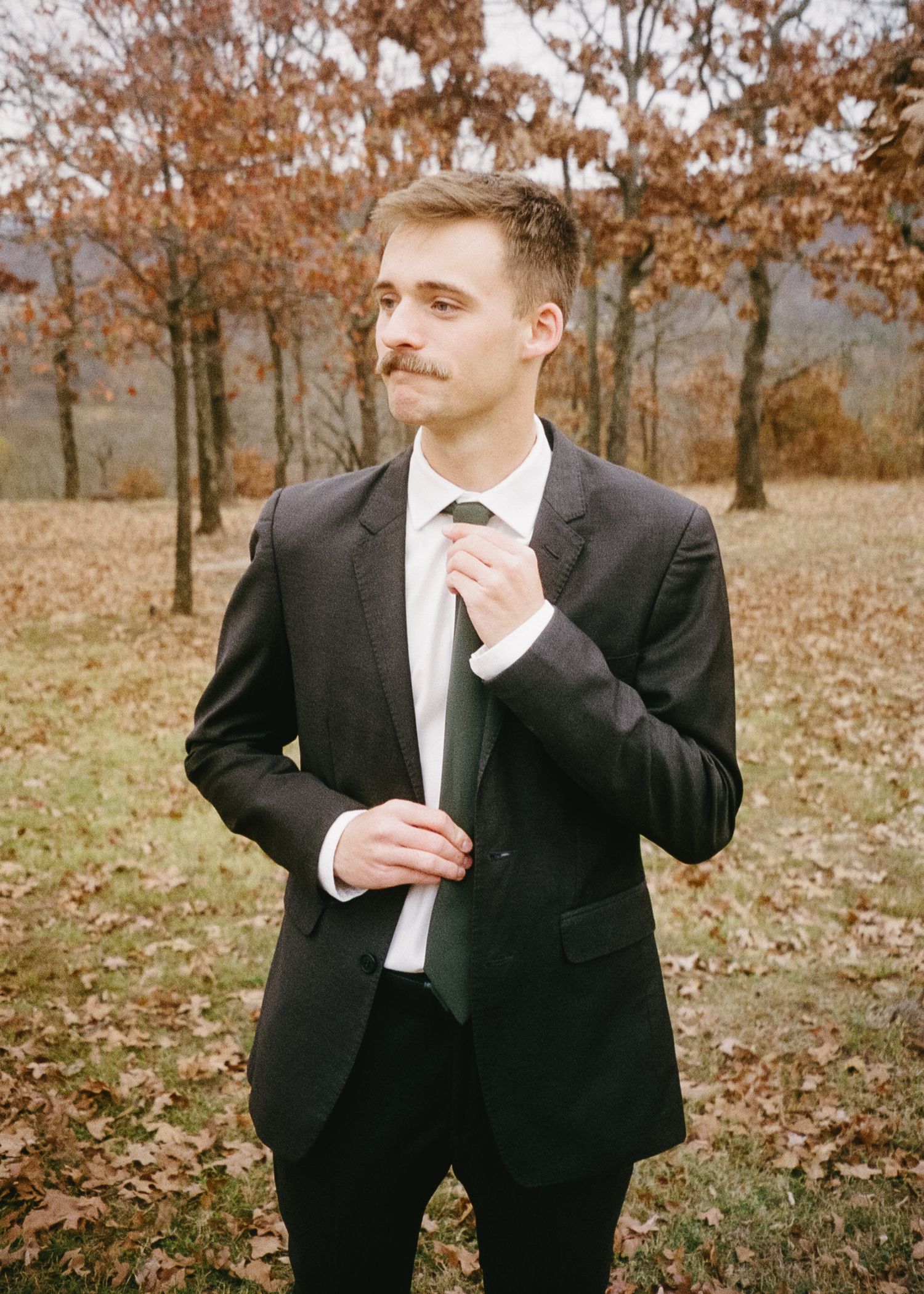 Man in black suit and white shirt posing playfully in an autumn field with fallen leaves.
