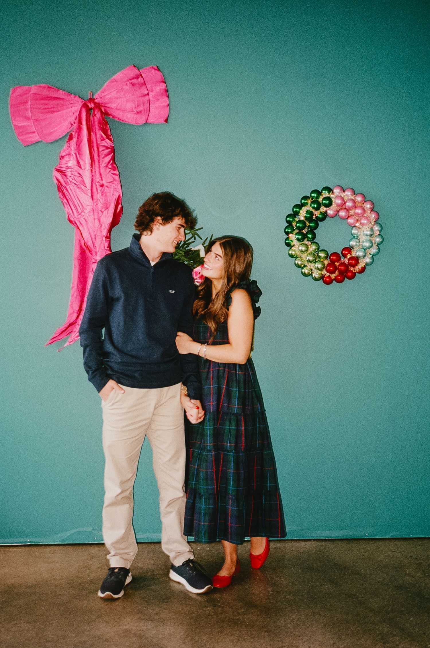 Two figures in a whimsical holiday photo with pink bows and mistletoe against a teal background.
