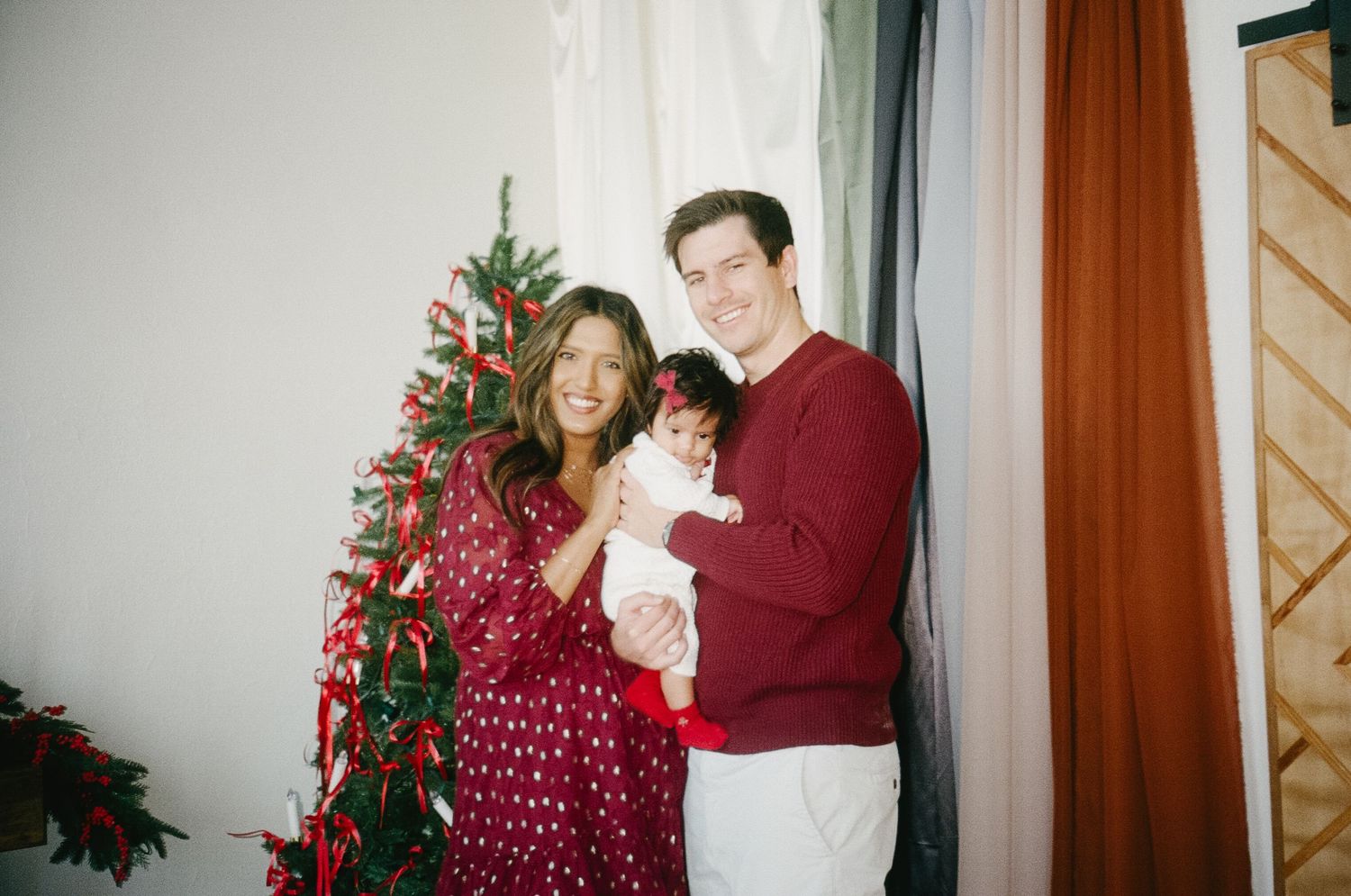 A couple in matching burgundy and white sweaters poses with their baby in front of a Christmas tree for holiday photos.