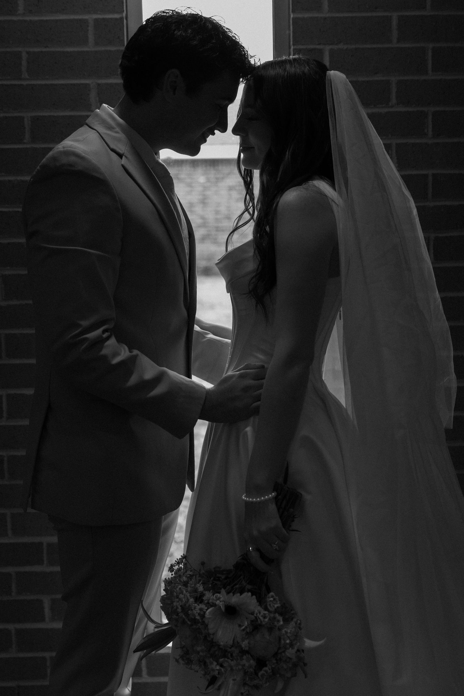Silhouetted wedding couple sharing an intimate moment against brick wall backdrop.
