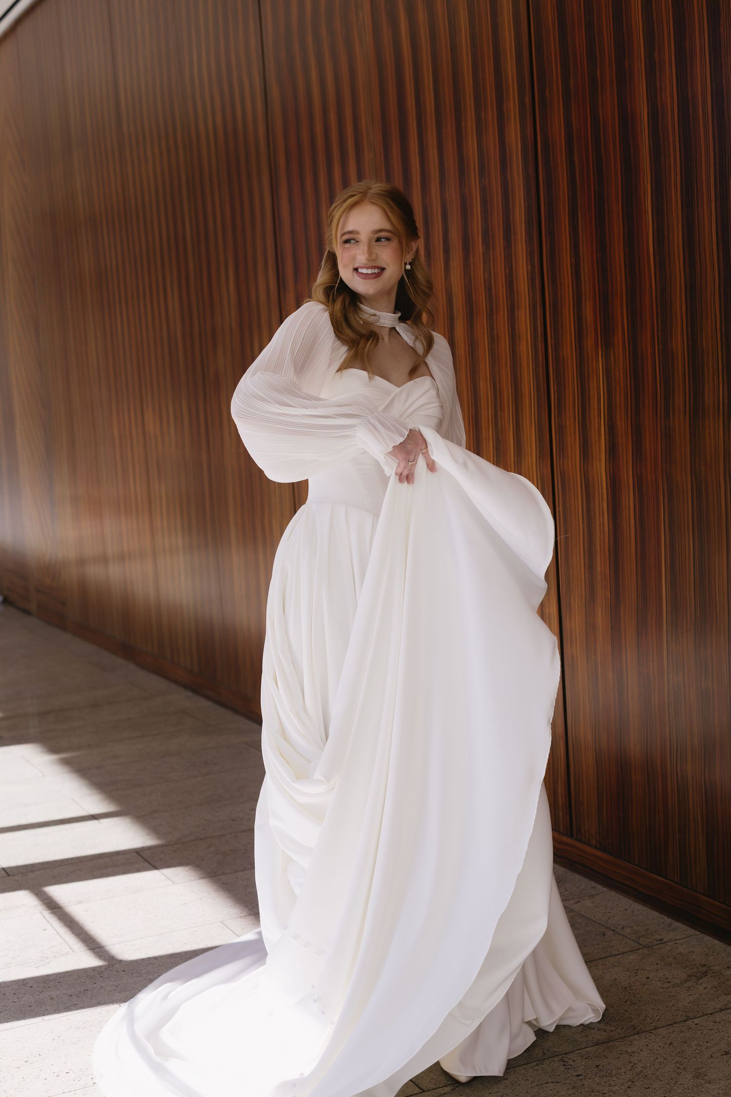 Flowing white wedding dress with dramatic sleeves photographed against wood paneling in sunlight.