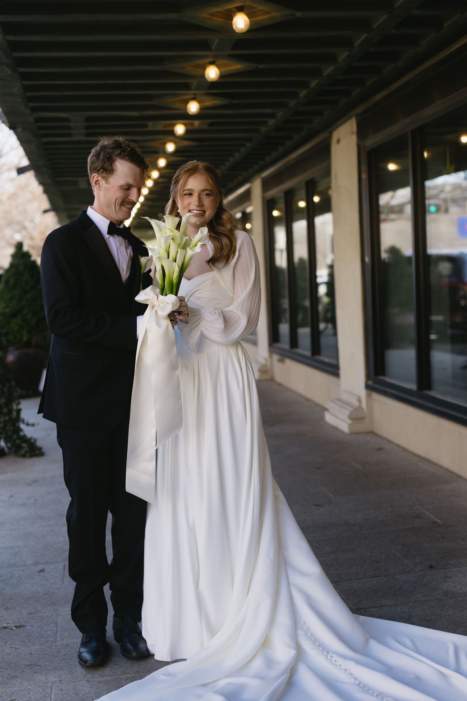 Wedding couple walks along a covered outdoor walkway with white dress and calla lily bouquet.