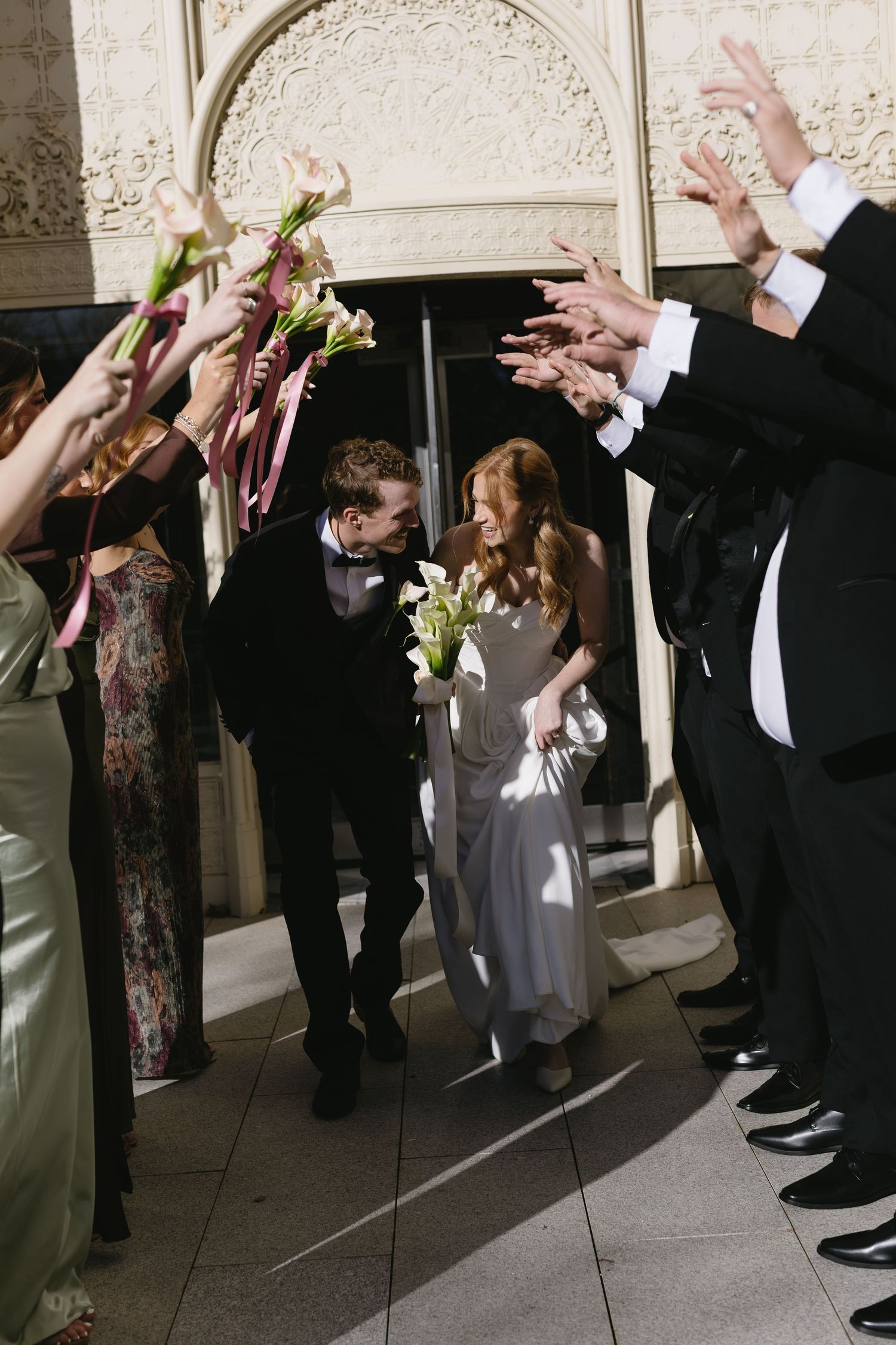Wedding party and guests raise their hands in celebration under an ornate archway.
