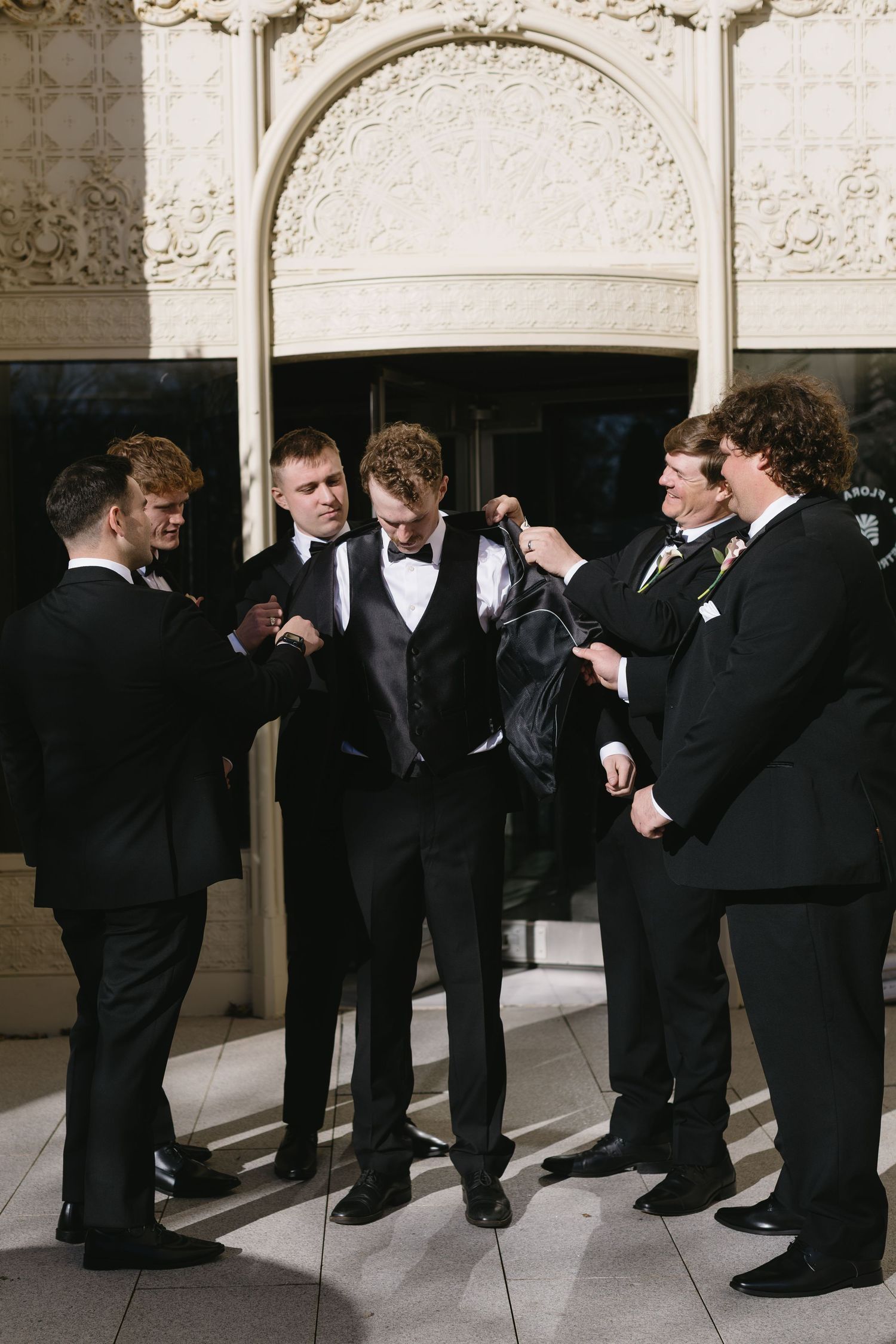Groomsmen in black tuxedos pose playfully in a line against an ornate wall.
