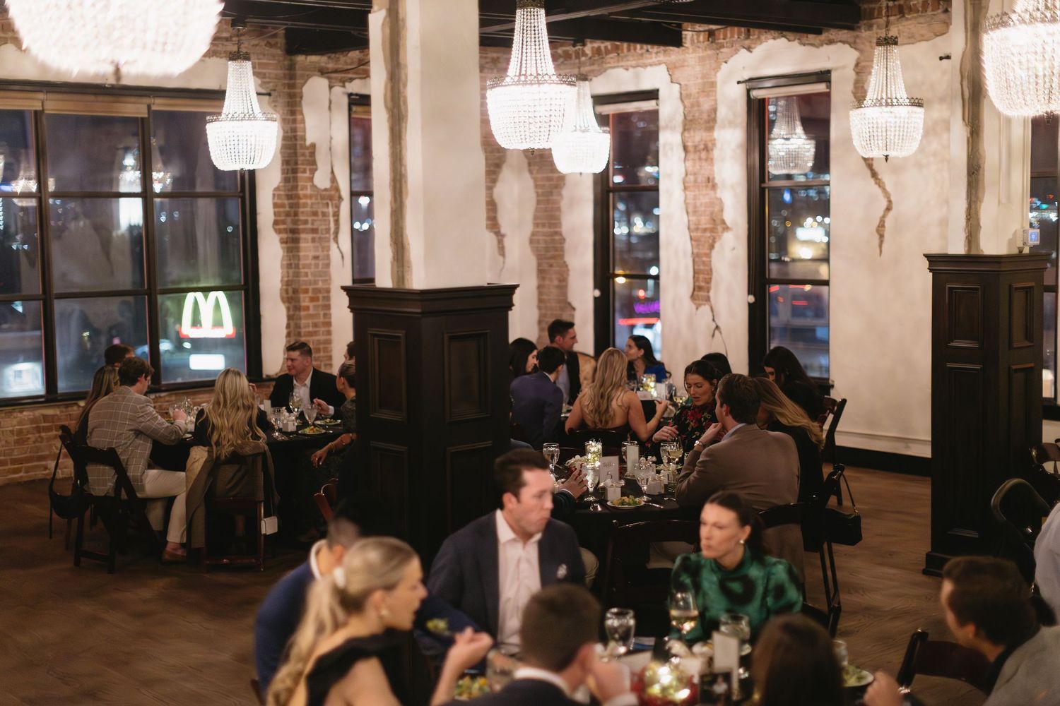 Crowd socializing in spacious restaurant with exposed brick walls and hanging chandeliers.