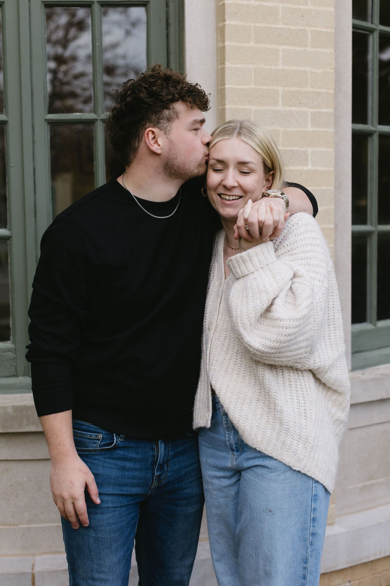 A couple in matching sweaters sharing an intimate moment outside a building with green trim.