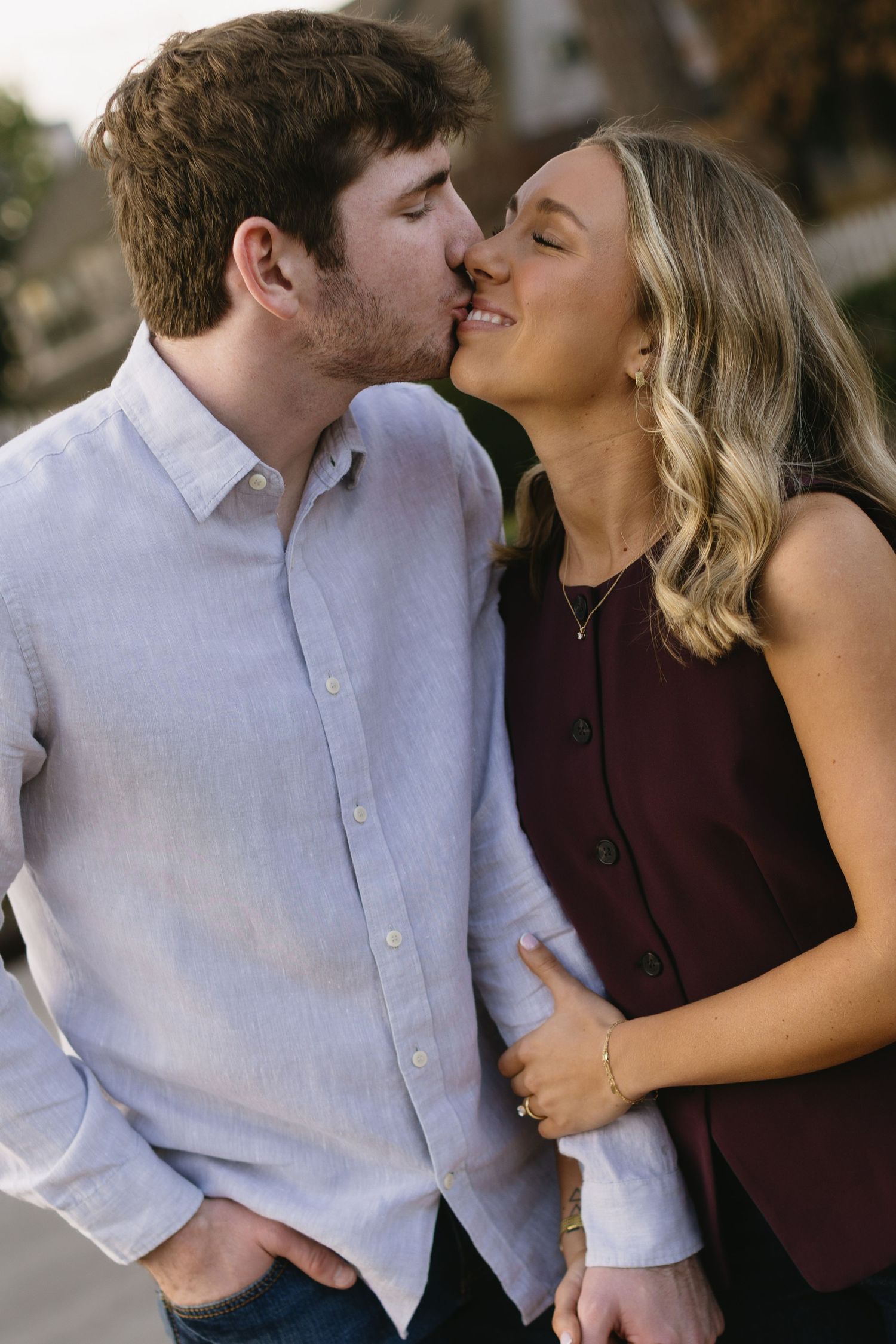 A casual couple sharing a kiss outdoors, with her in burgundy and him in light blue.