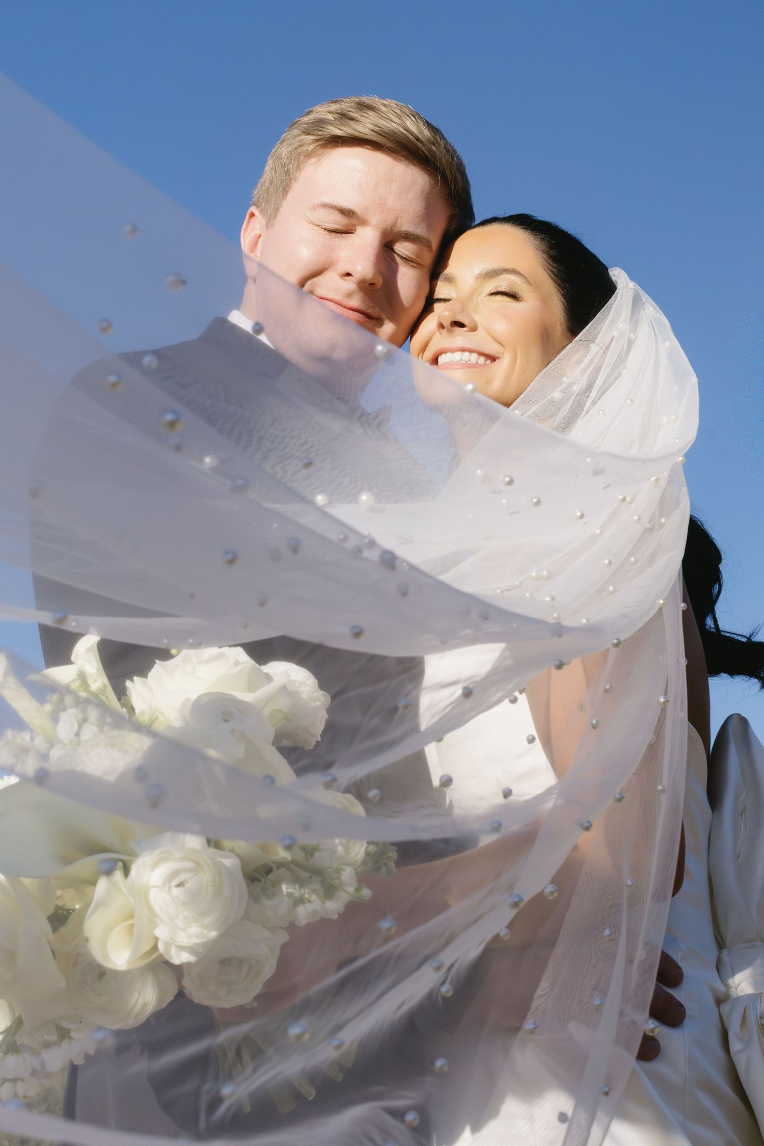 Delicate white veil flows in the breeze against bright blue sky during outdoor wedding photography.