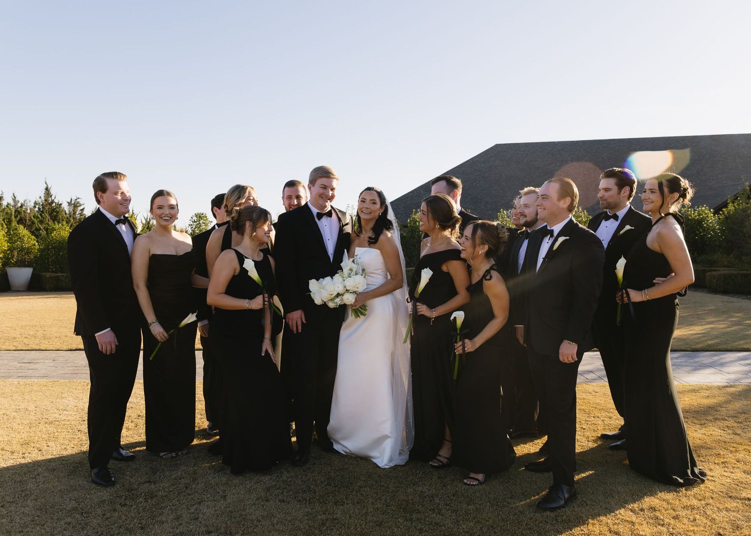 Wedding party in black attire poses together on a grassy hill during golden hour.