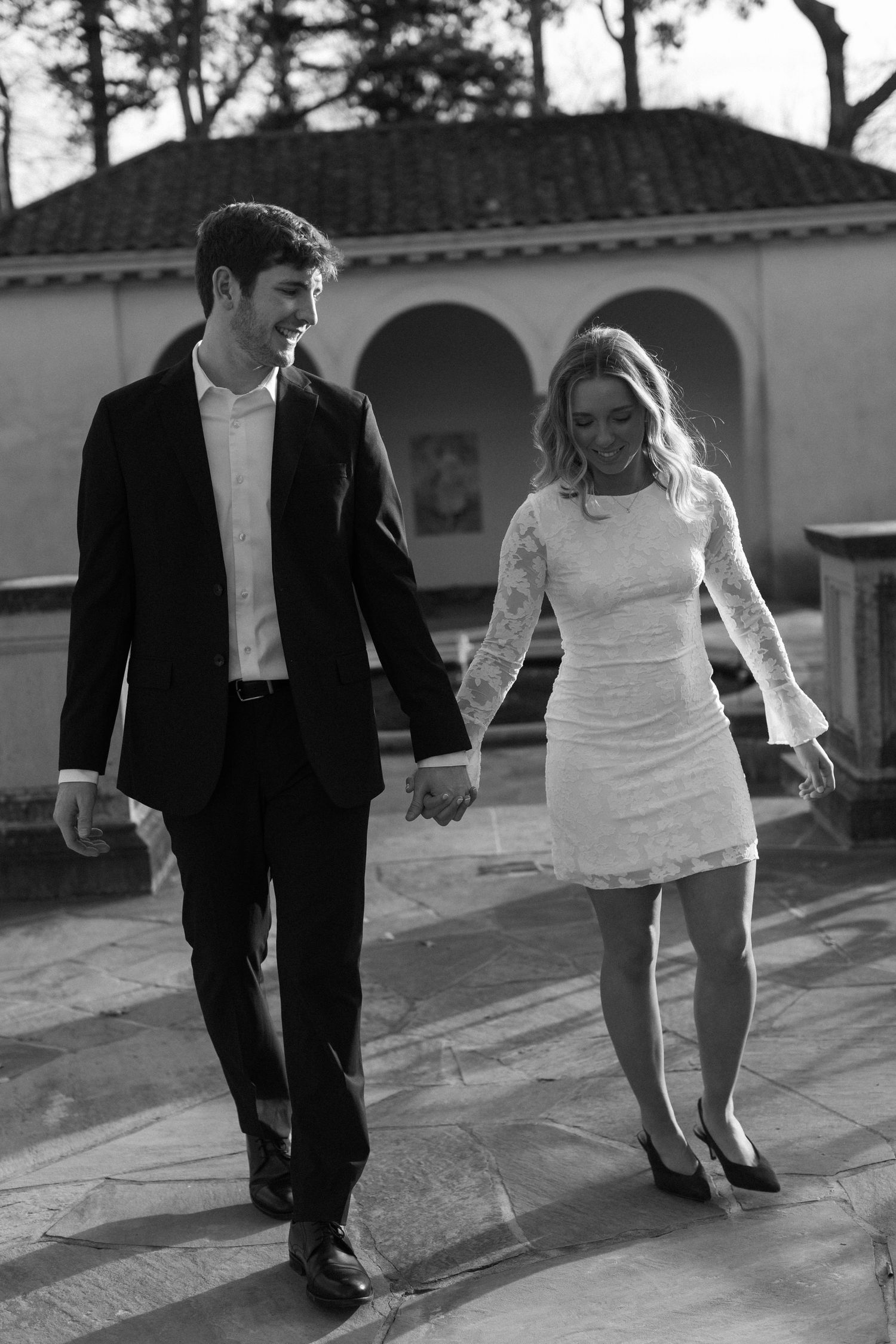 A couple walks playfully together outdoors in formal attire during a black and white photoshoot.