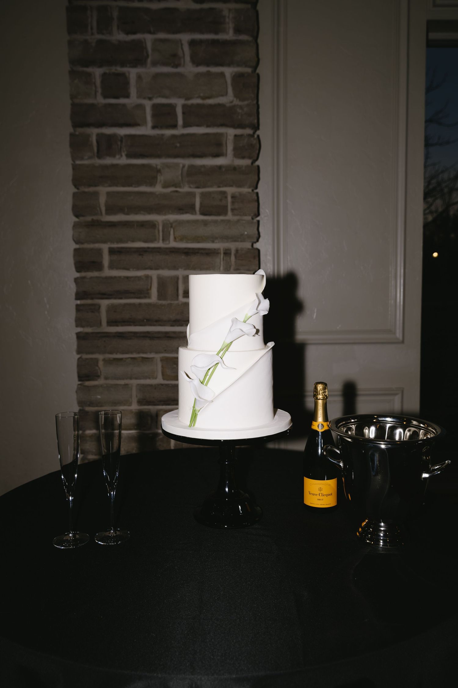 Minimalist white wedding cake displayed on dark table with champagne bottles.