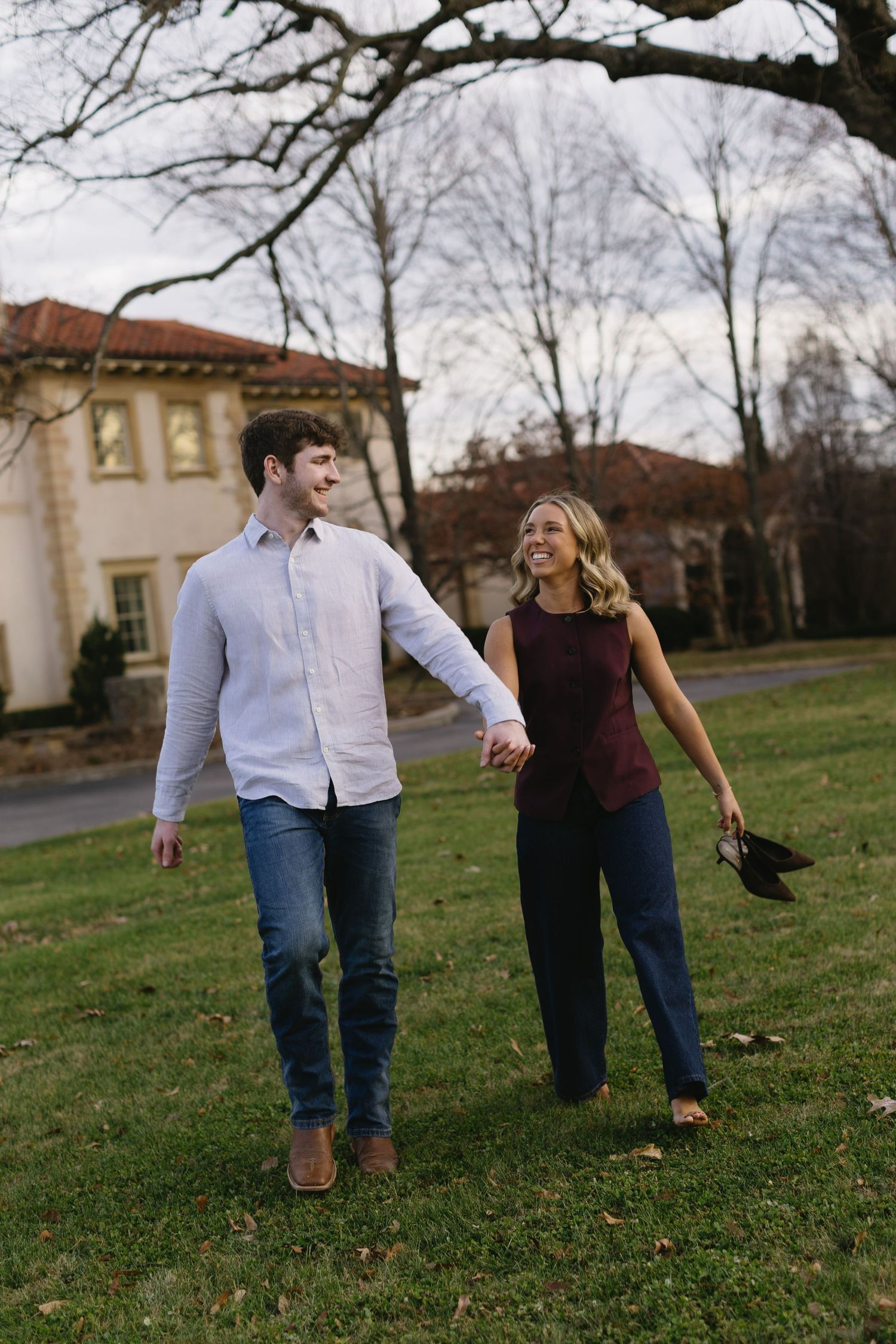 A couple walks hand in hand across a lawn with historic buildings in the background.