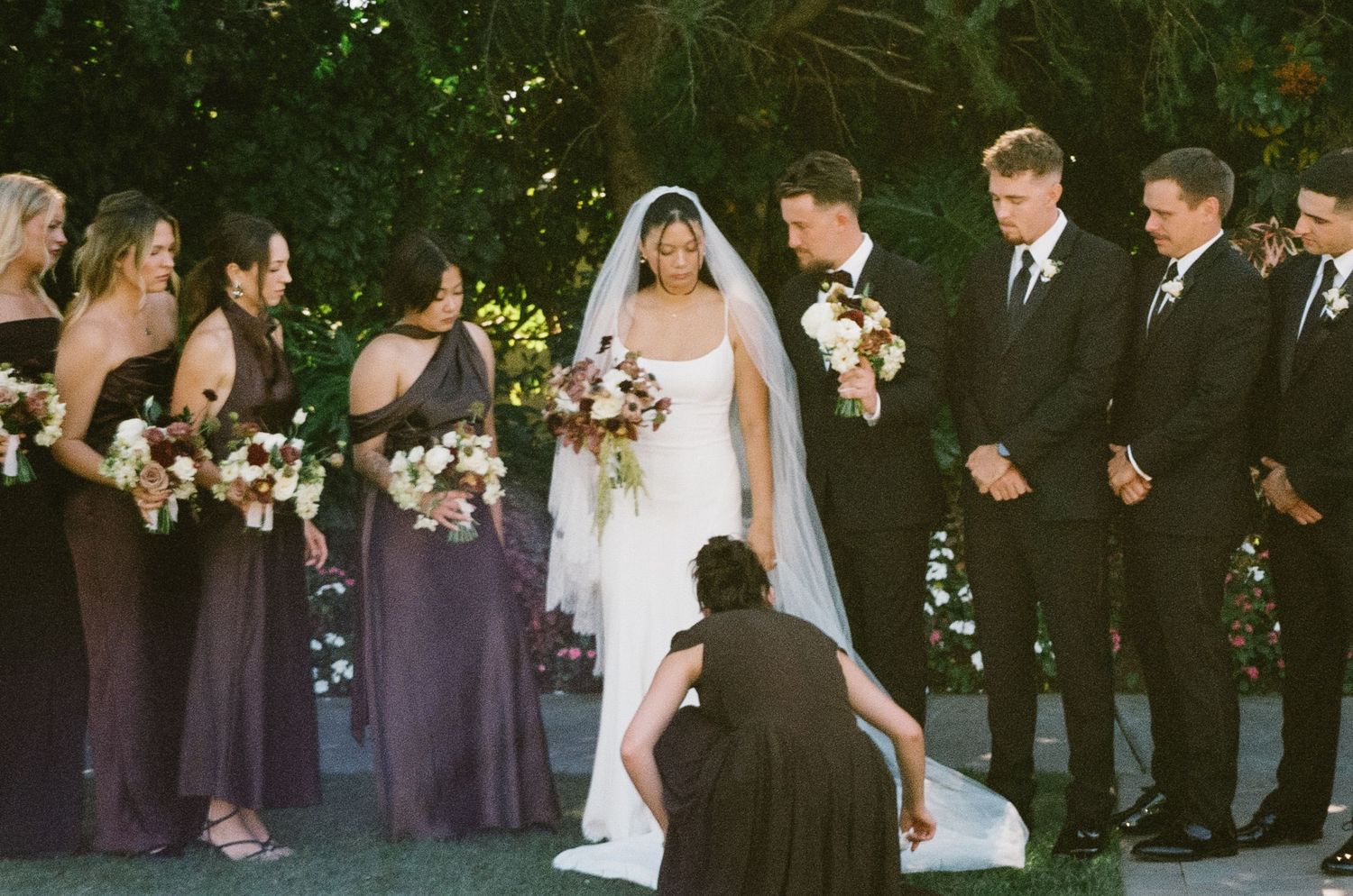 Wedding party members in purple dresses and dark suits stand together outdoors during a formal ceremony.