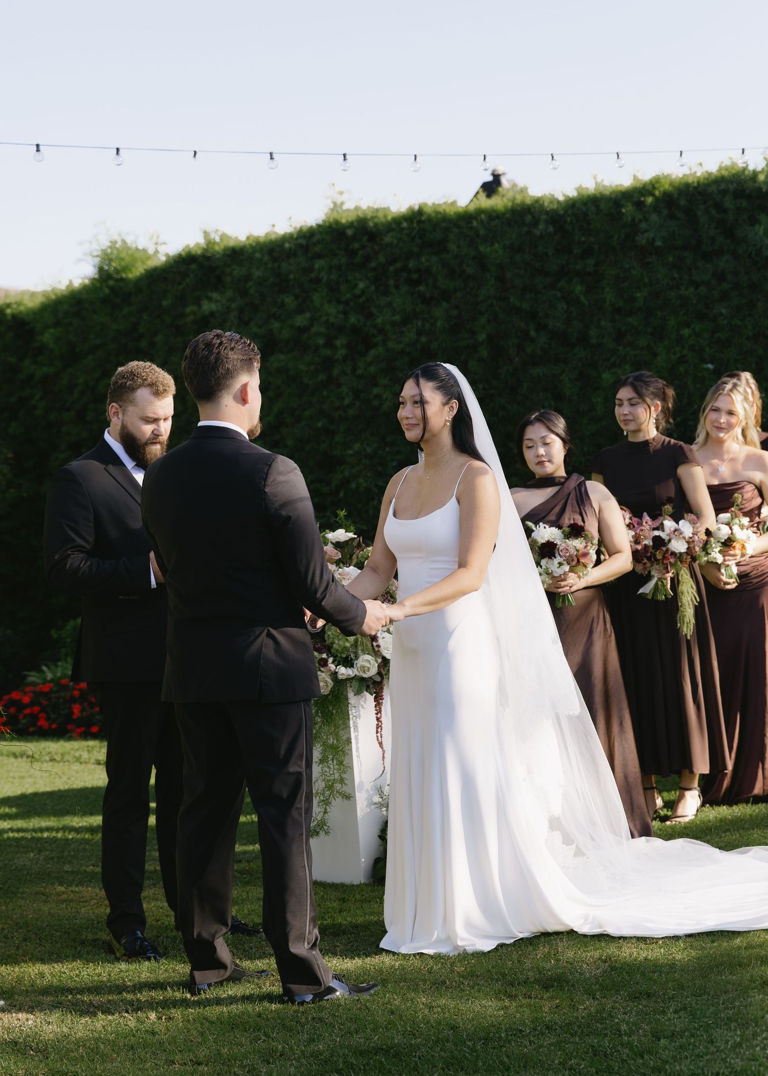 Wedding party in brown and black attire gathers outdoors with string lights and hedges in the background.