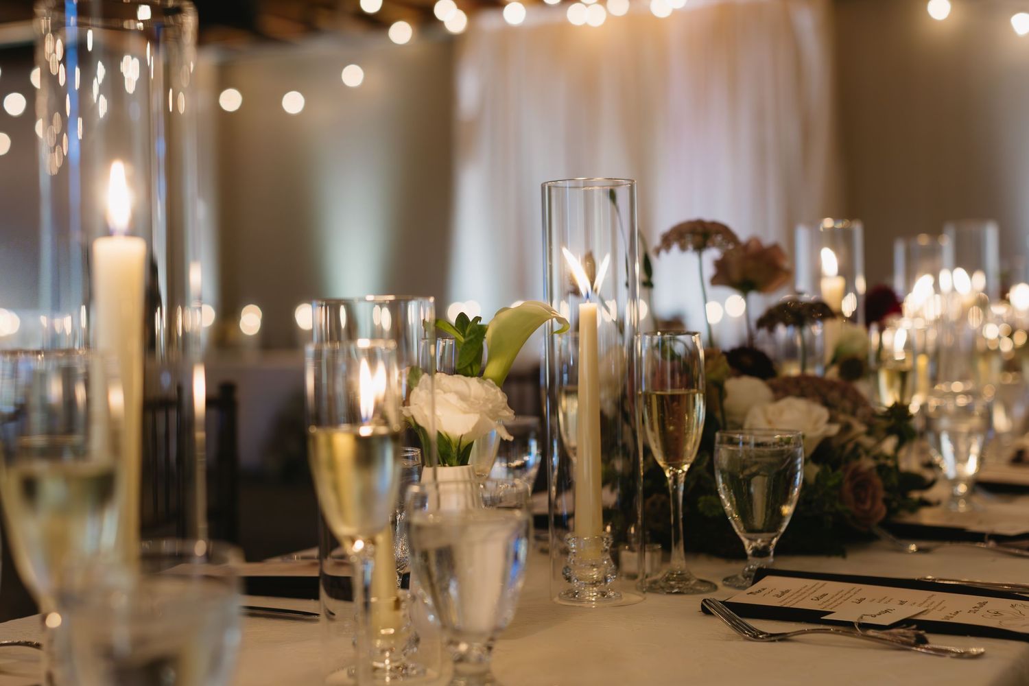 String lights illuminate an elegant wedding reception table setting with crystal glasses and white floral centerpieces.