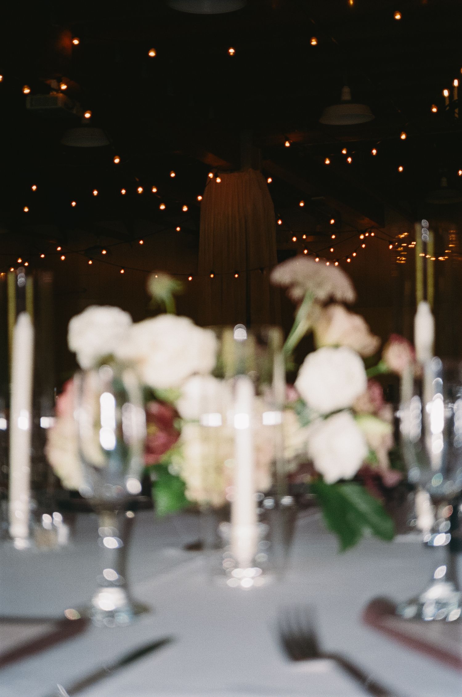 Elegant wedding reception table setting with white floral centerpieces and twinkling lights overhead in a dark venue.