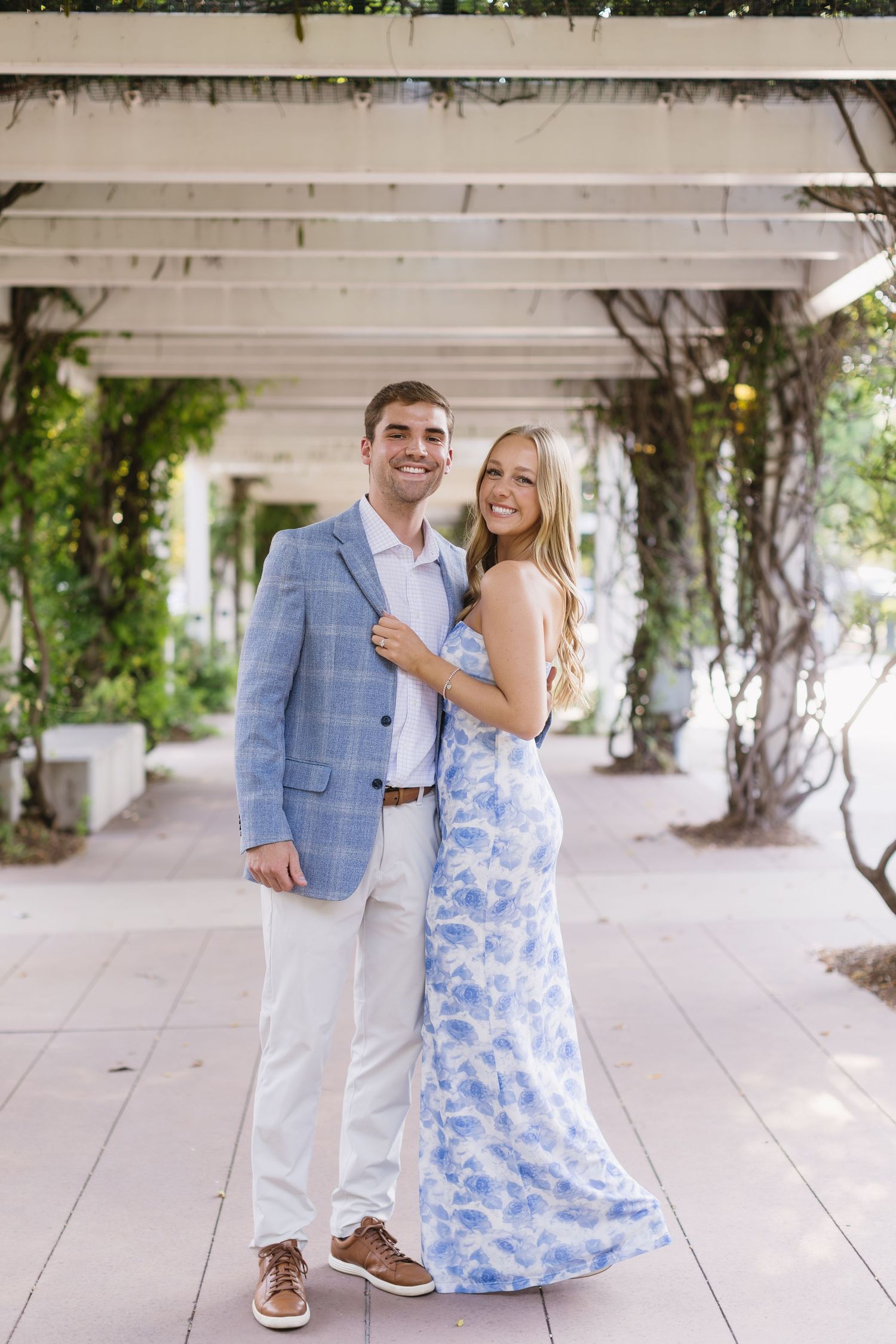 A couple poses in formal attire on a covered walkway with vines, wearing blue and white coordinated outfits.