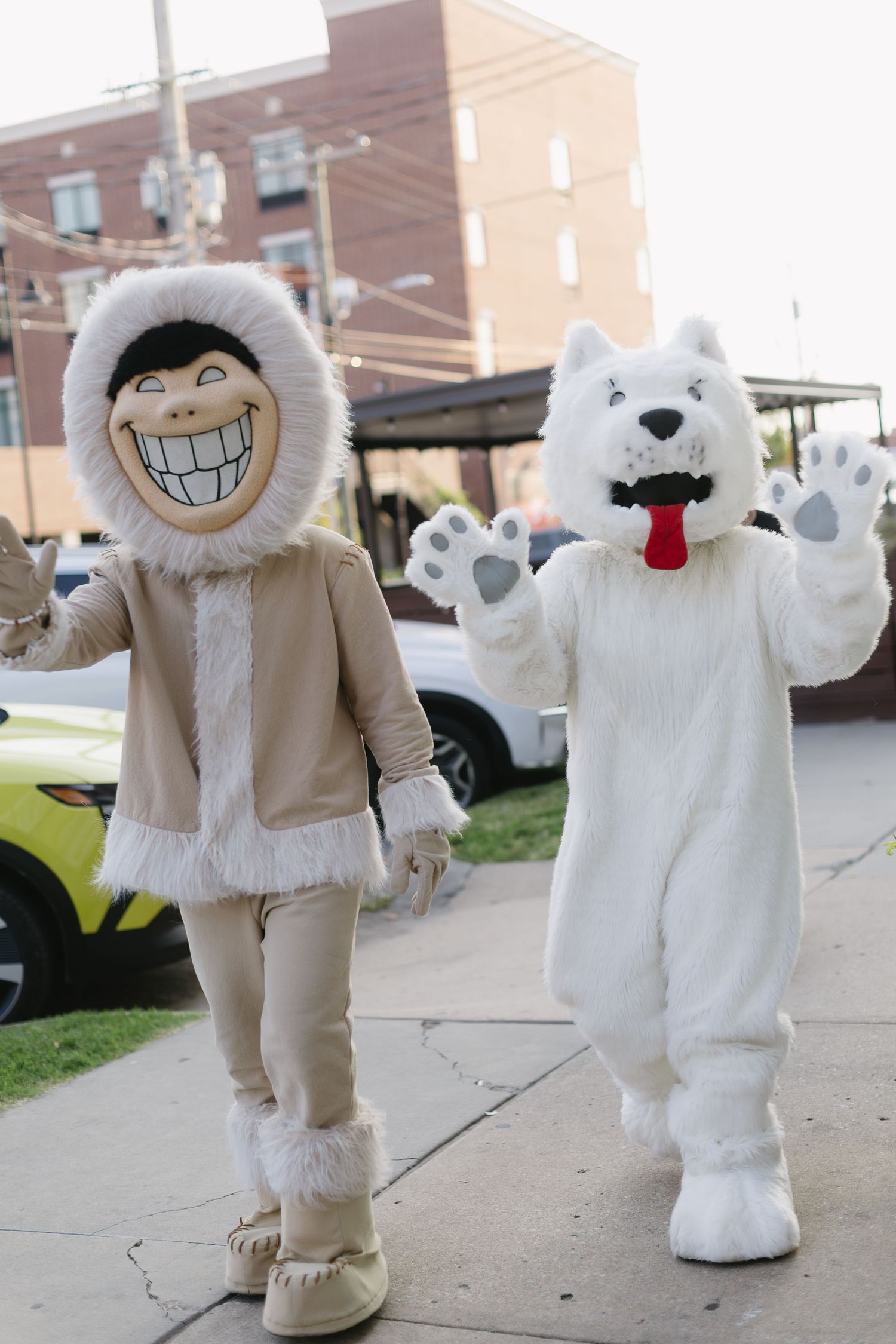 Two mascot costumes in furry white and beige outfits with big smiles posing outdoors on a sidewalk.