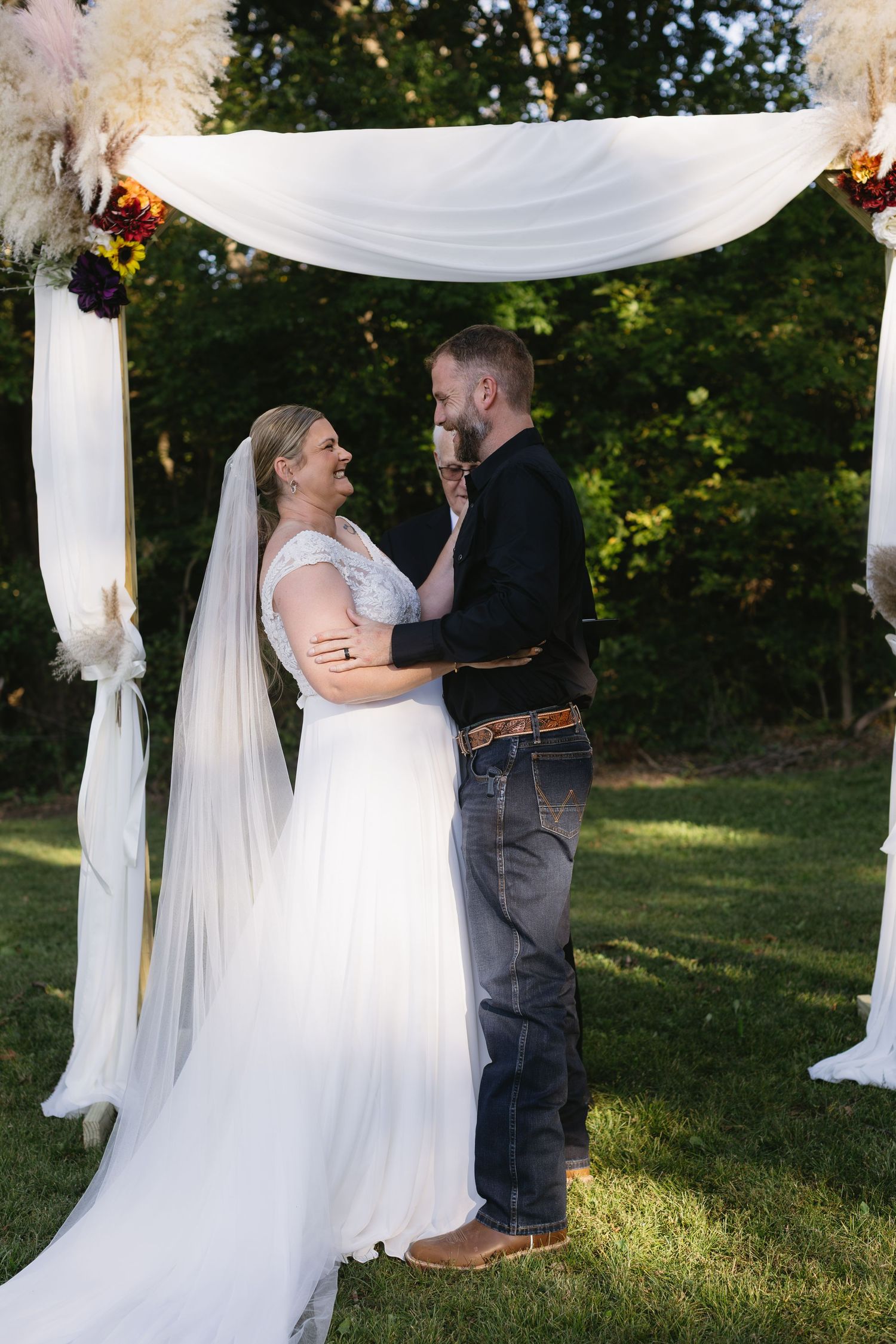 Romantic outdoor wedding ceremony under white fabric draped archway in natural sunlight.