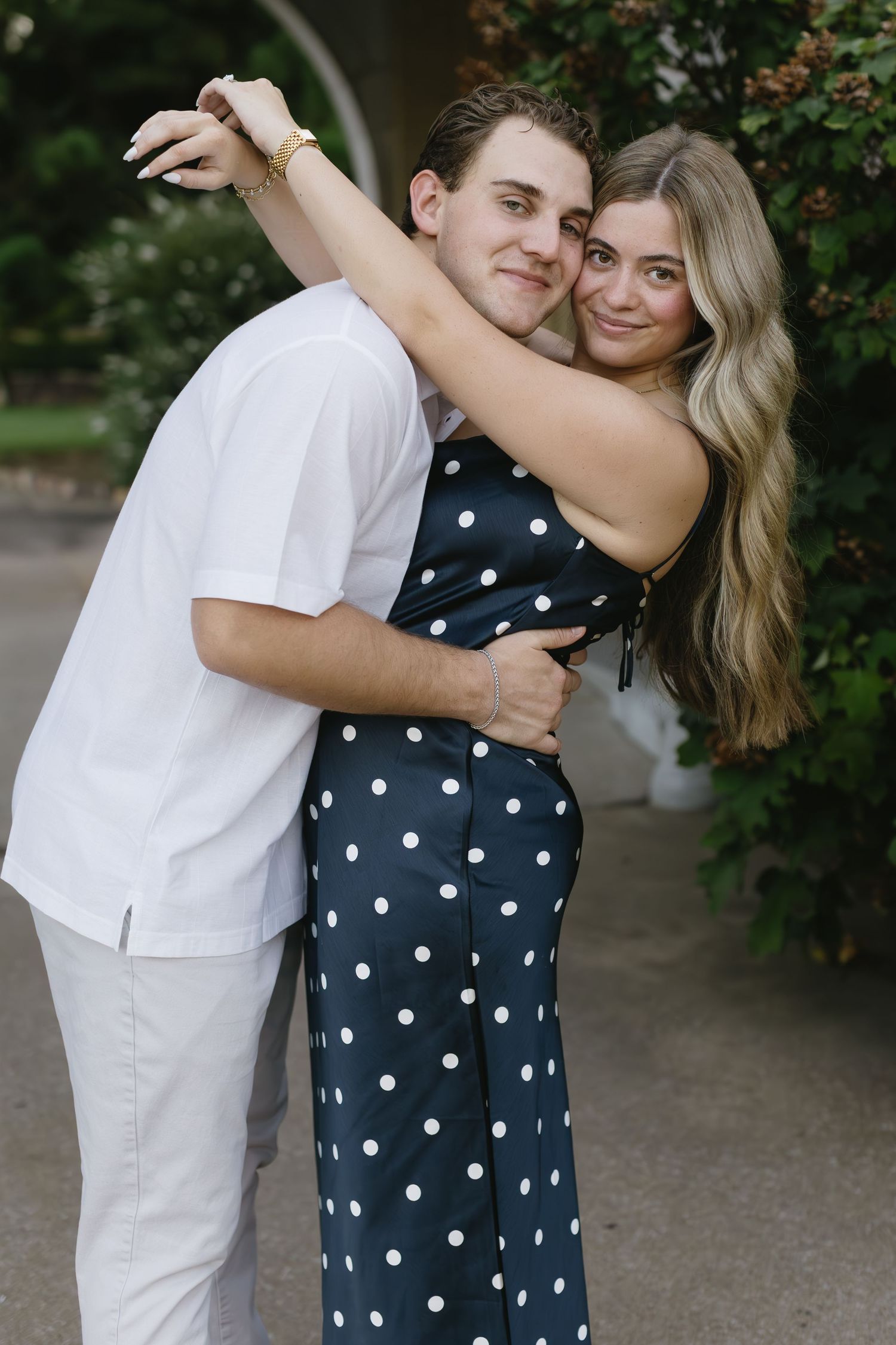A couple embraces in a romantic pose outdoors, with one wearing white and the other in a navy polka dot dress.