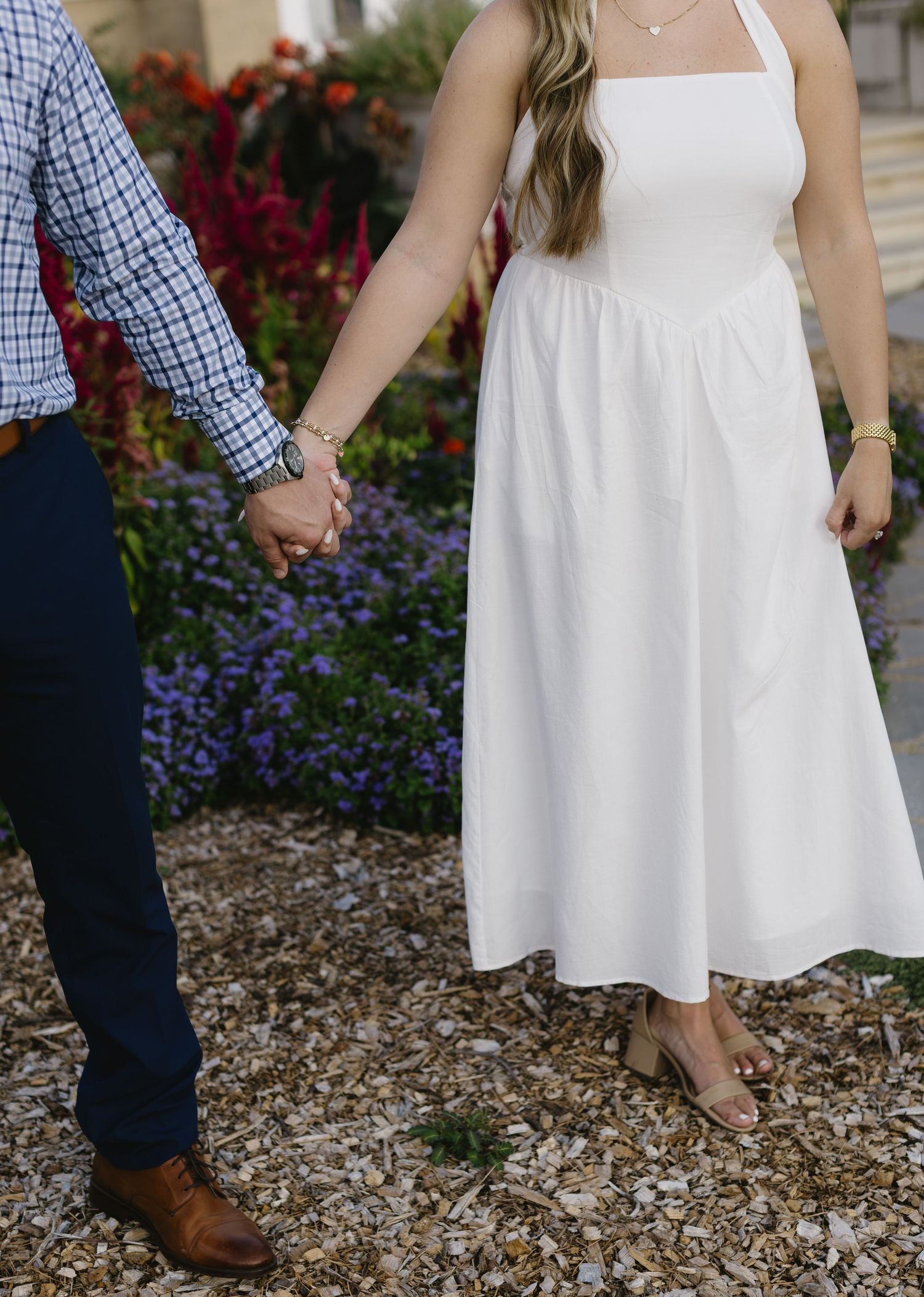 Close-up of couple holding hands while standing in a garden, showing white dress and navy pants details.
