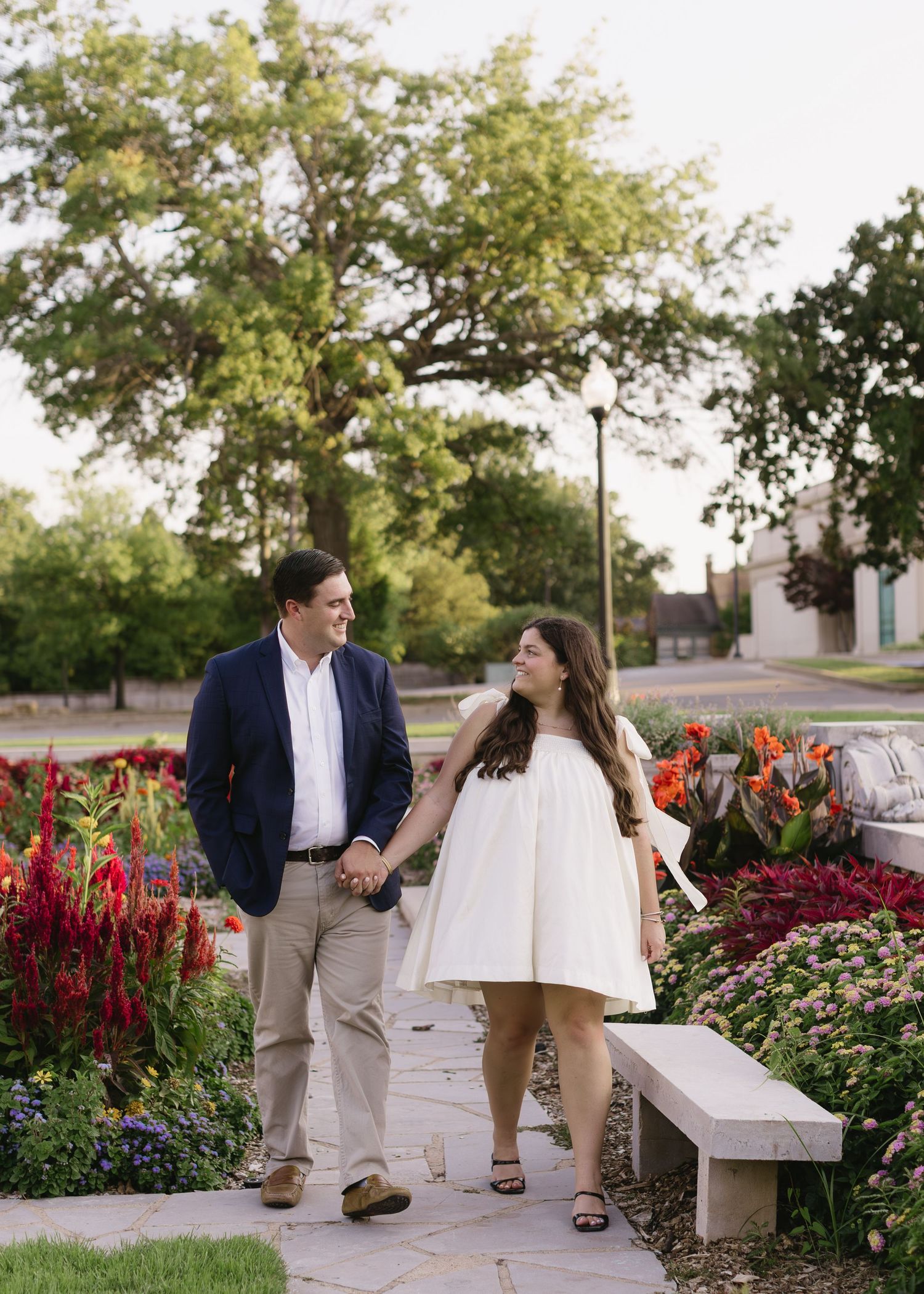 A couple walks hand in hand through a garden with red flowers and trees.