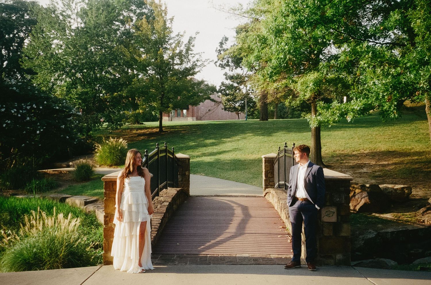 A couple stands together on a winding garden path surrounded by lush greenery and trees in an outdoor setting.
