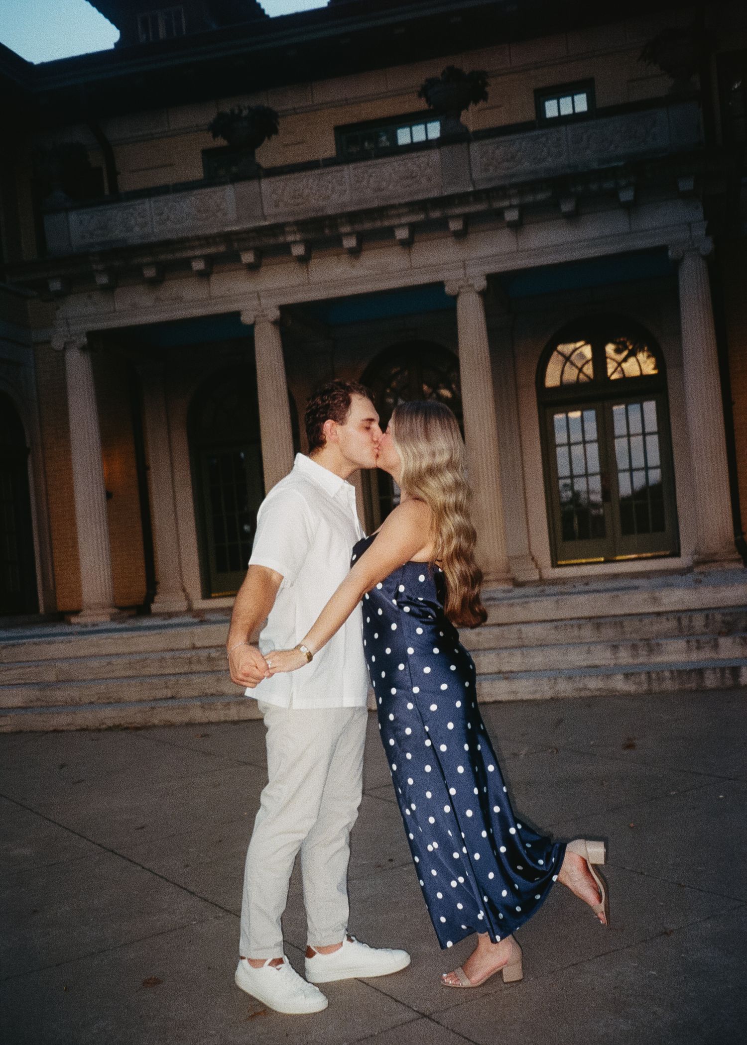 A couple in white and polka dot outfits dances together in front of a historic building at dusk.