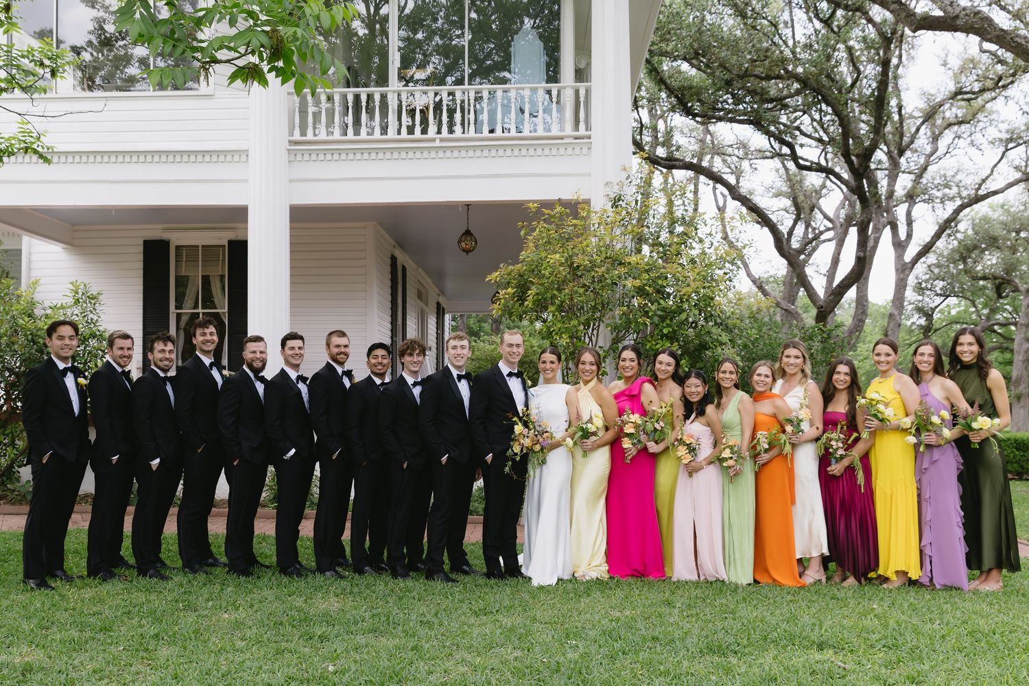 A wedding party poses outdoors in colorful dresses and suits along a grand white columned porch.