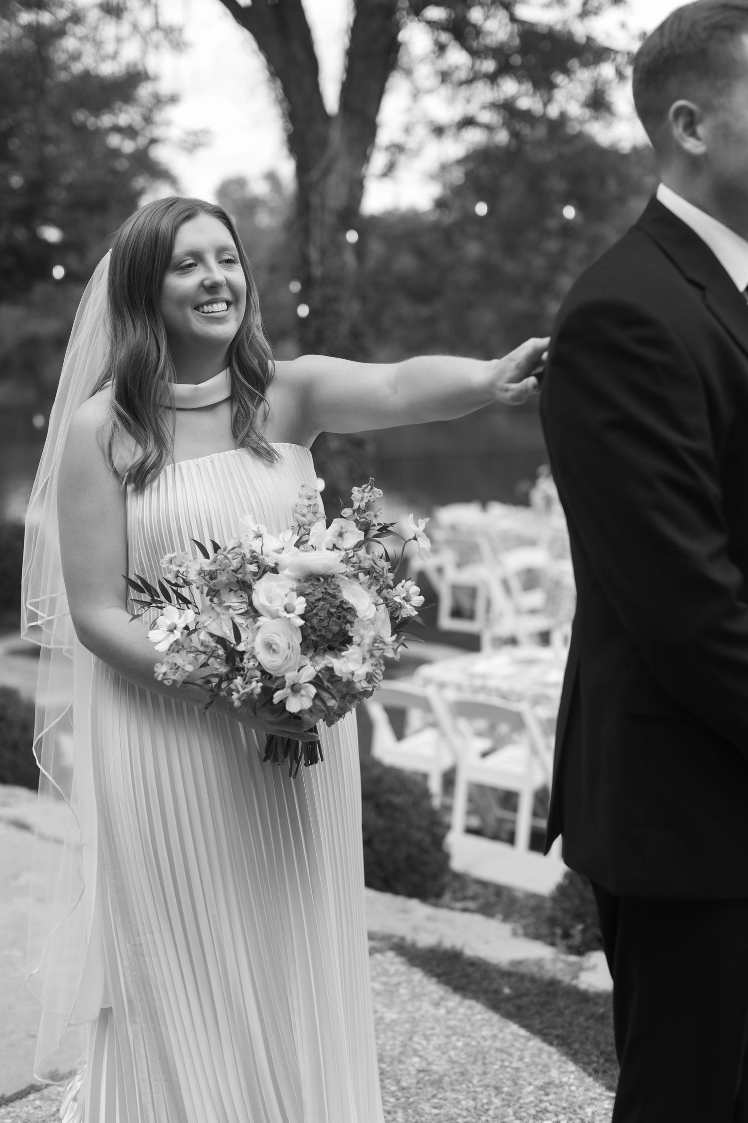 Elegant outdoor wedding scene with white pleated dress and floral bouquet in black and white.