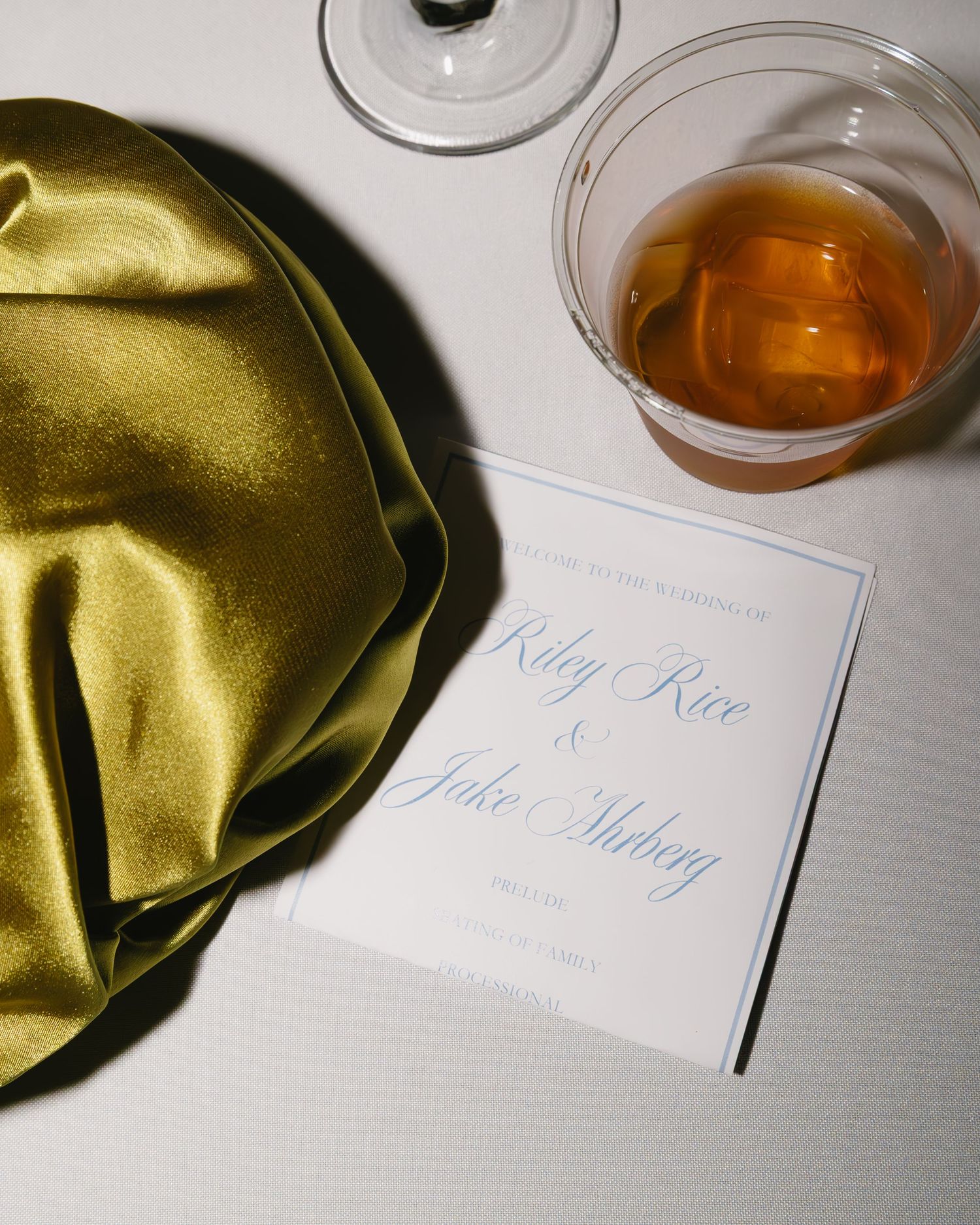Elegant place cards and gold napkins are arranged with amber-colored drinks on a white surface.