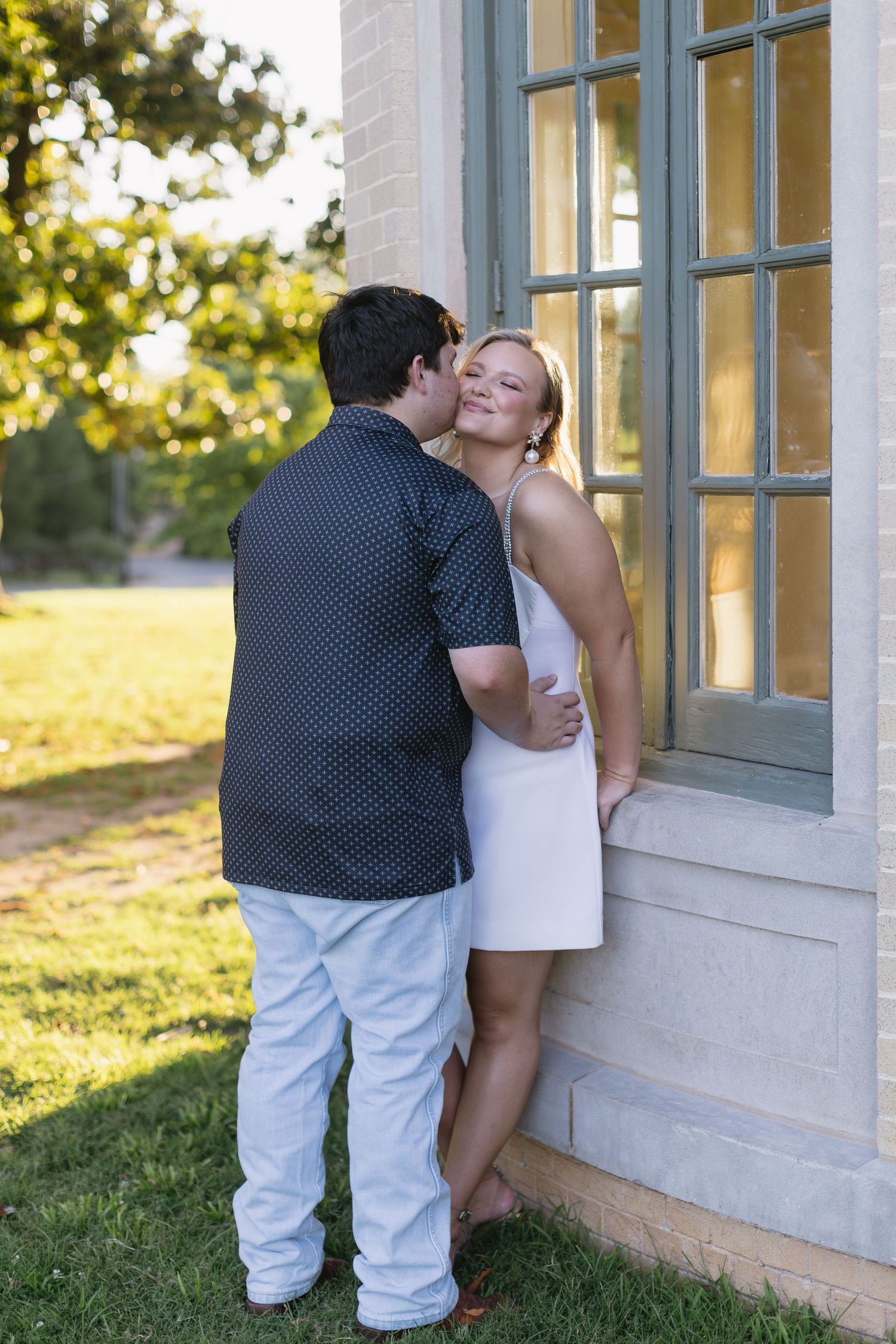 A couple shares an intimate moment against a building exterior with windows in golden sunlight.