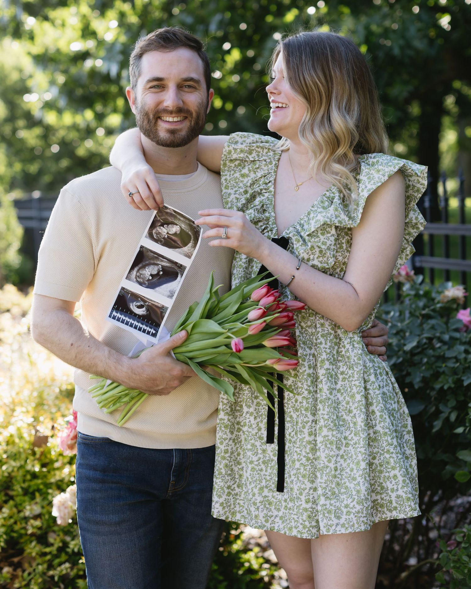 Couple celebrates with tulip flowers and ultrasound photos in a sunny garden setting.