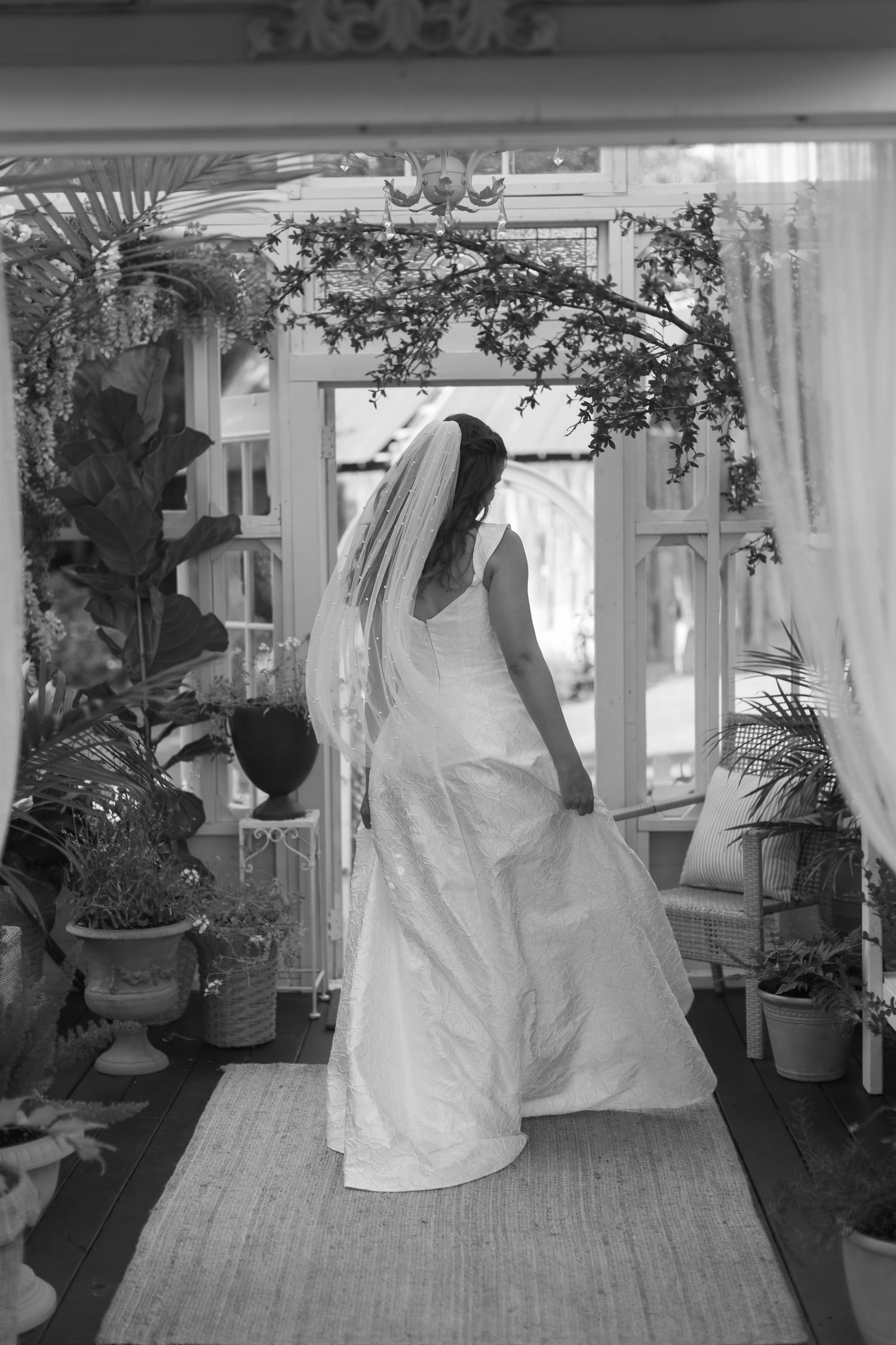 A bride in a white gown stands in a greenhouse surrounded by potted plants and natural light.