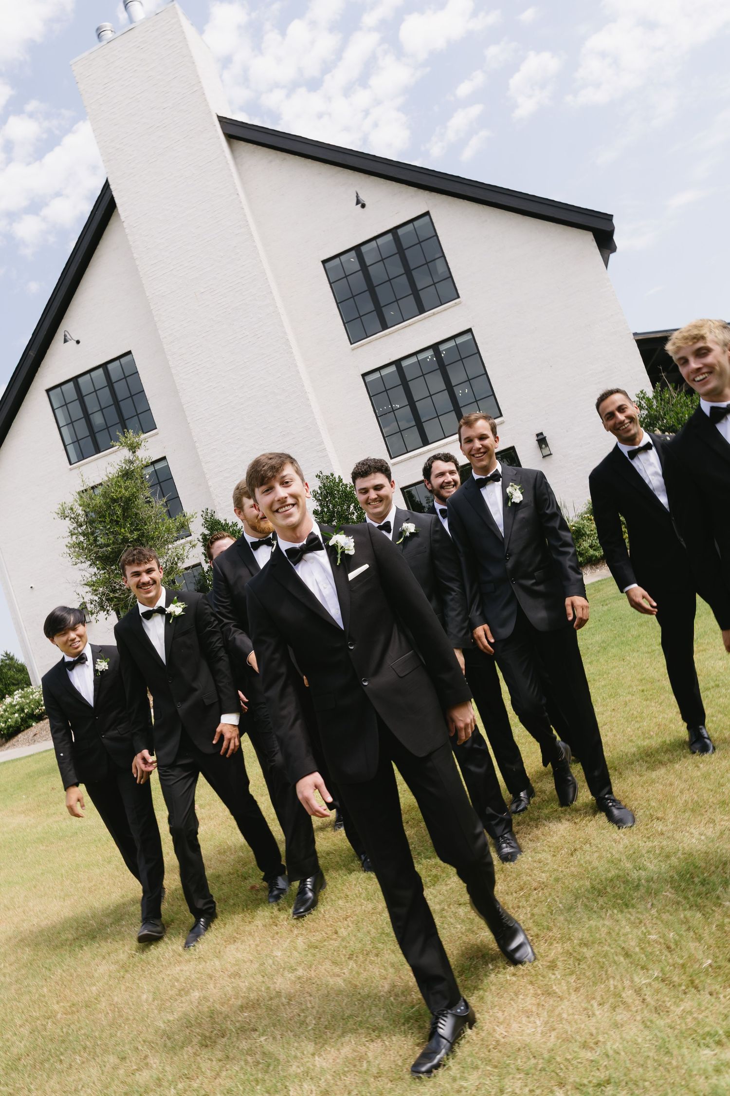 A group of groomsmen in black tuxedos pose together on green grass outside a modern white building.