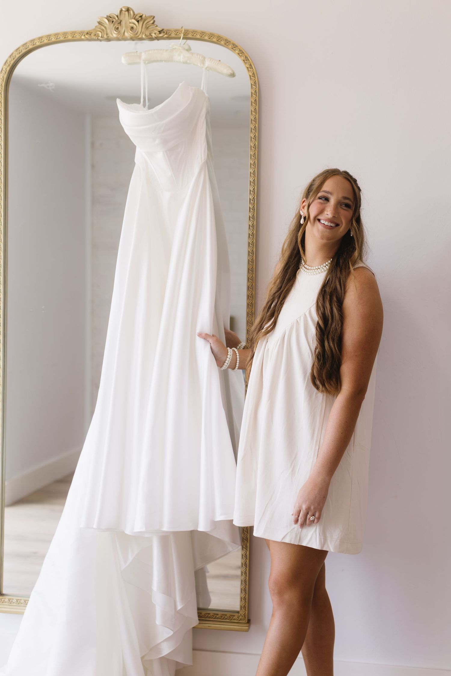 White wedding dress hanging in front of ornate gold-framed mirror.