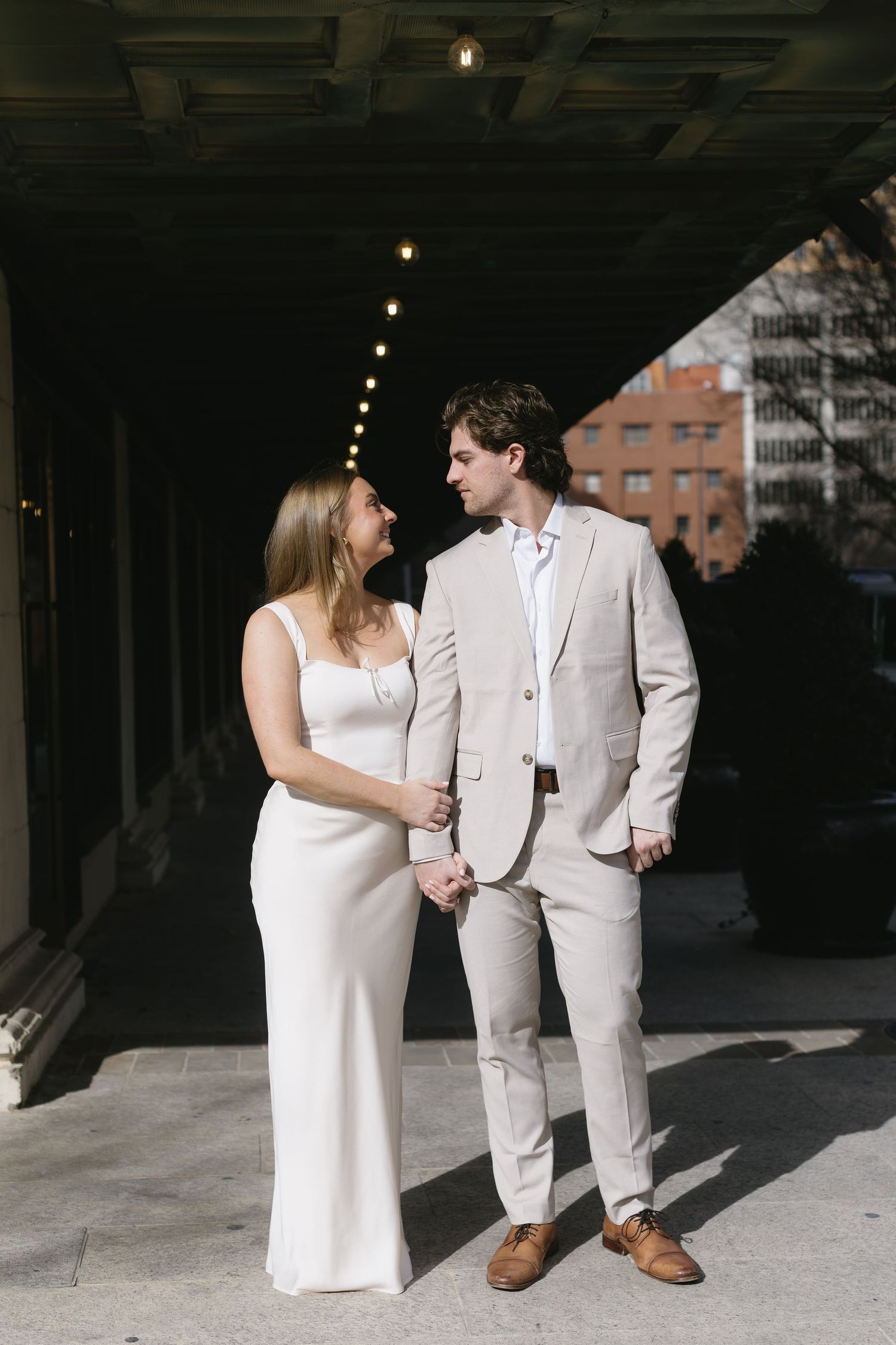 A couple in matching light-colored formal wear stands together under an industrial ceiling.