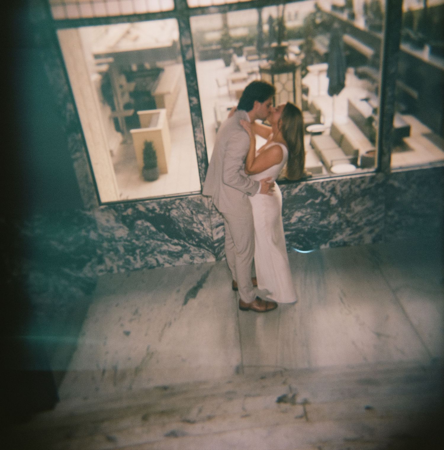 A romantic couple embraces in formal wear by a window in vintage-style photography.