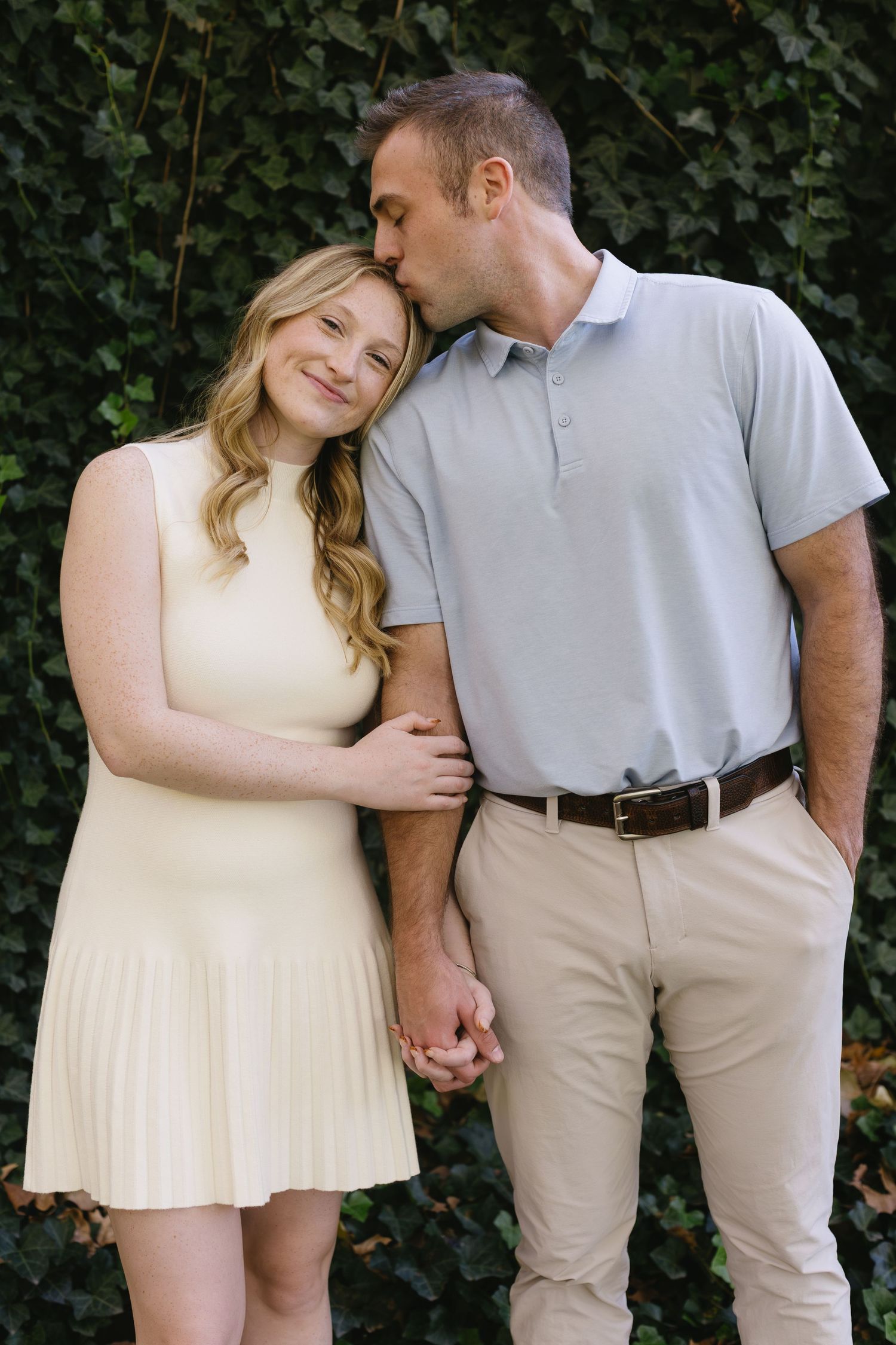 A couple in light-colored outfits shares an intimate moment against an ivy wall.