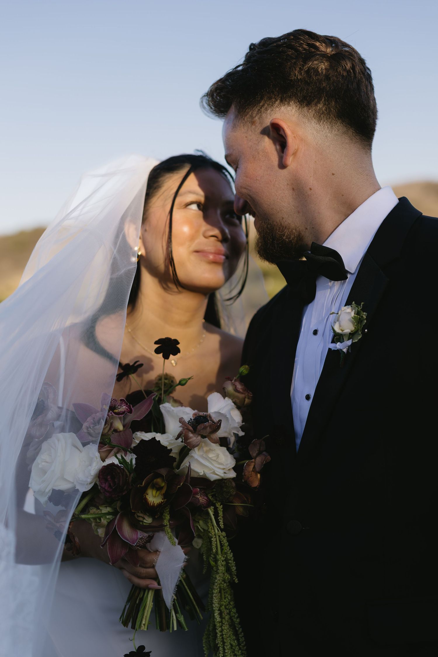 Romantic sunset silhouette of newlyweds with burgundy bouquet and flowing veil.