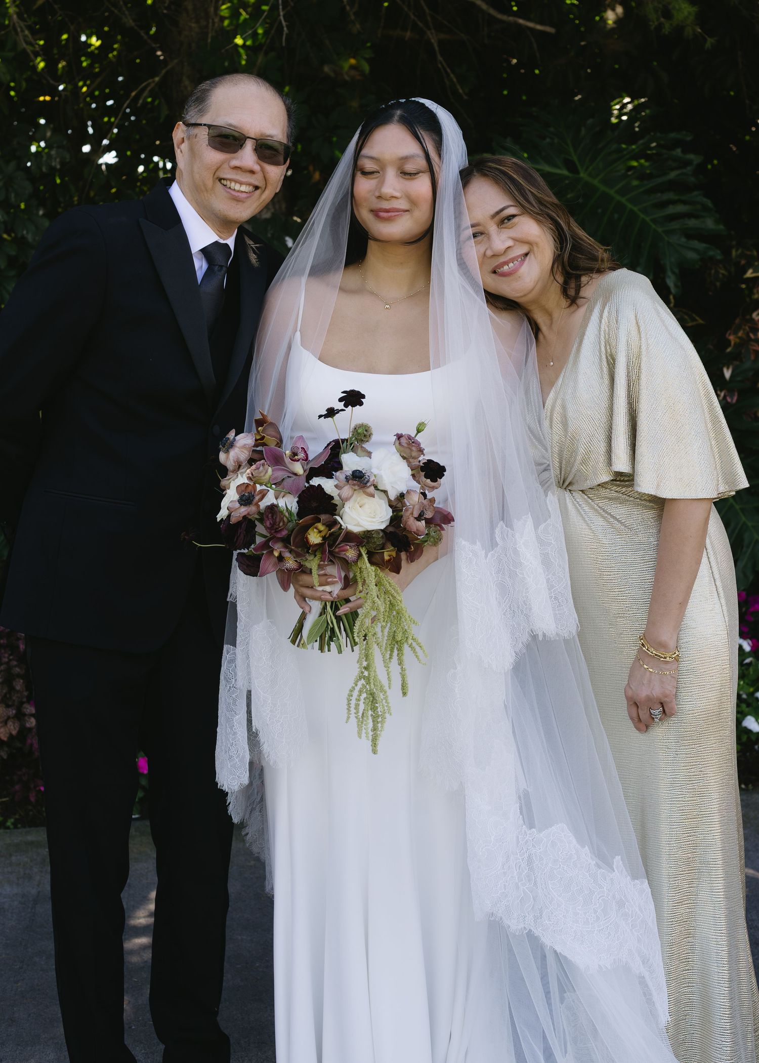 A bride in a white dress holds a burgundy floral bouquet while posing with guests outdoors.