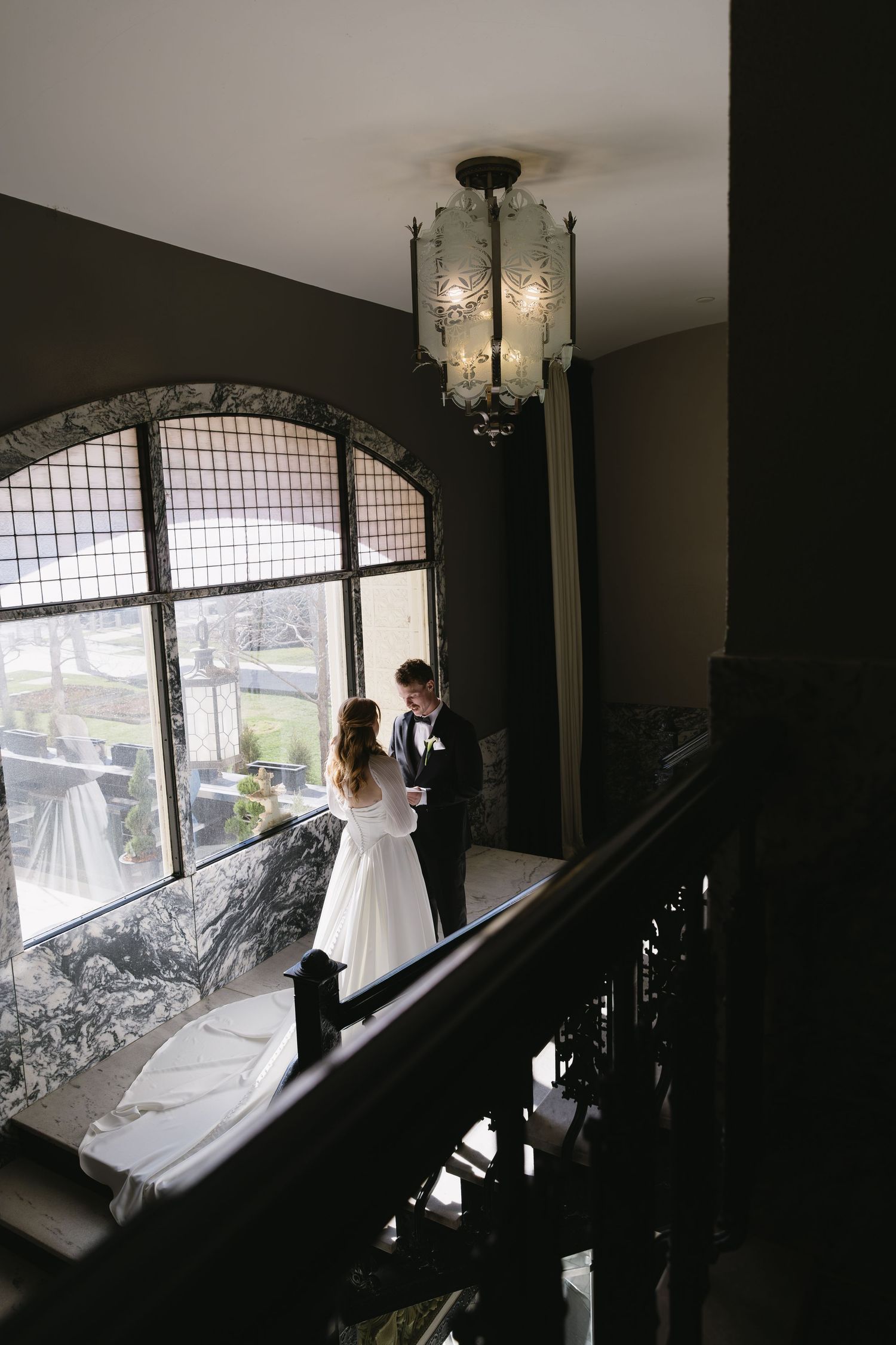 Wedding couple silhouetted against large windows in an elegant staircase setting.