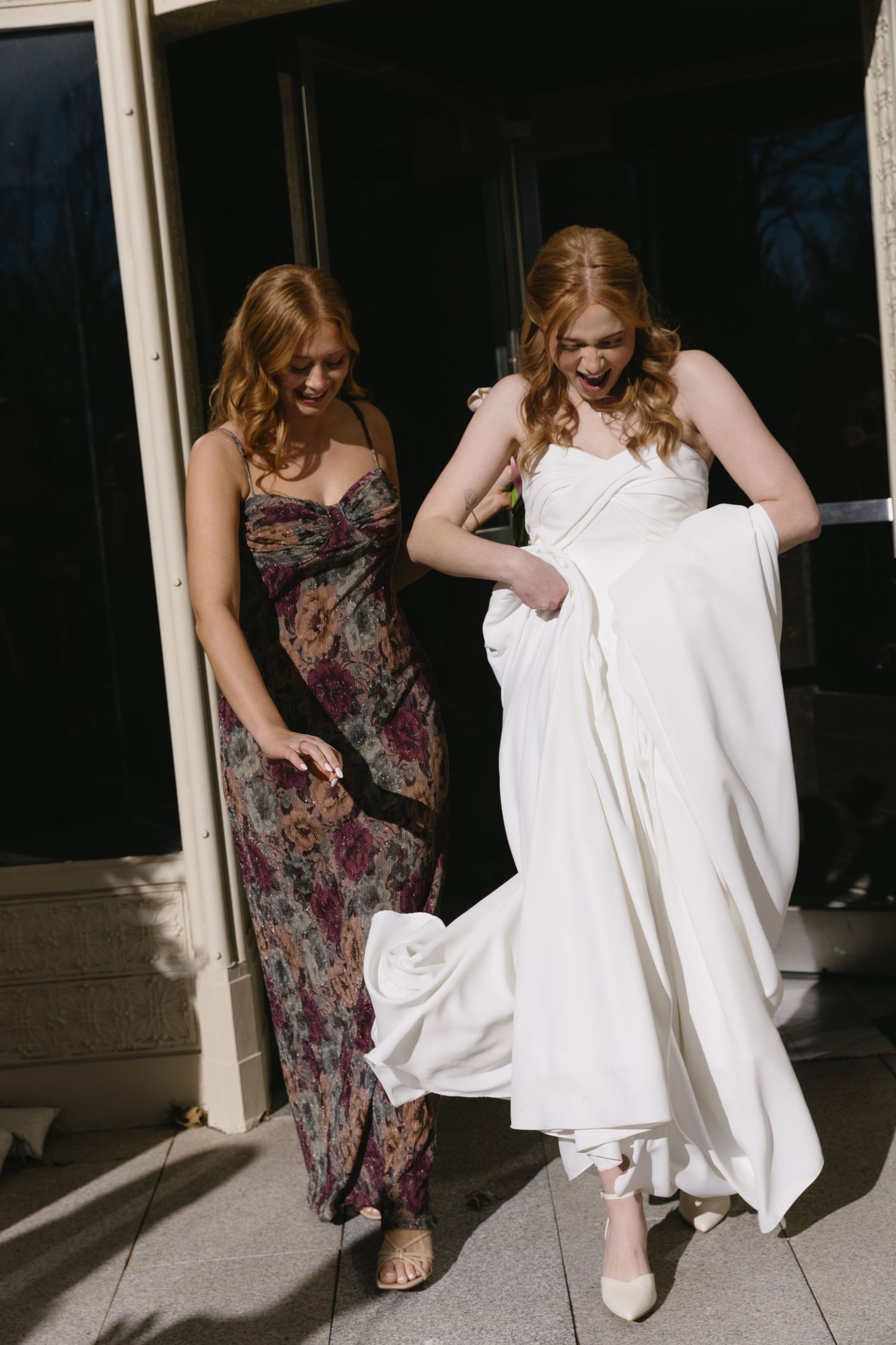 Two women walk together, one in a floral dress helping another in a white wedding gown with her dress.